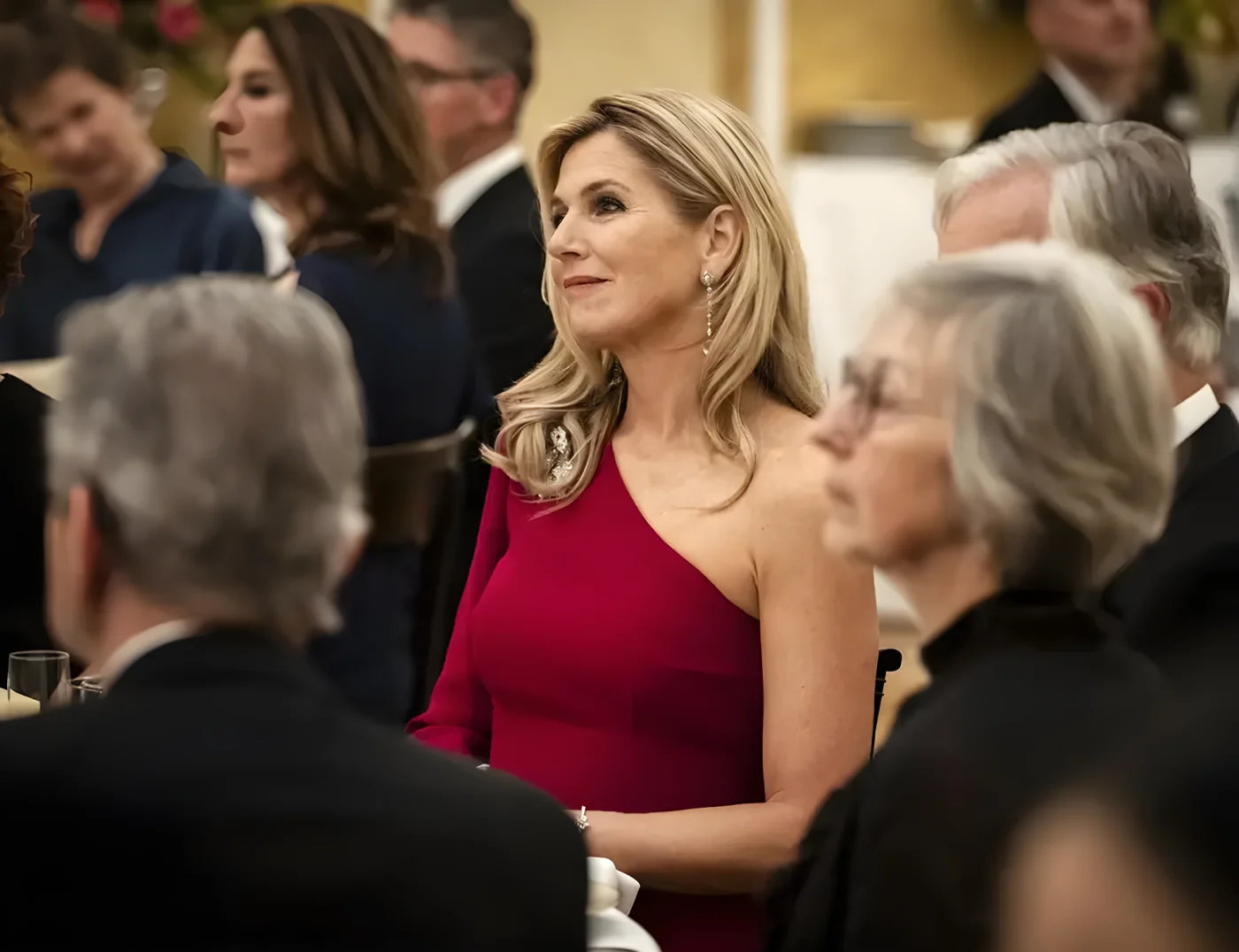 Queen Máxima of the Netherlands in a red dress at a formal event, surrounded by other guests.