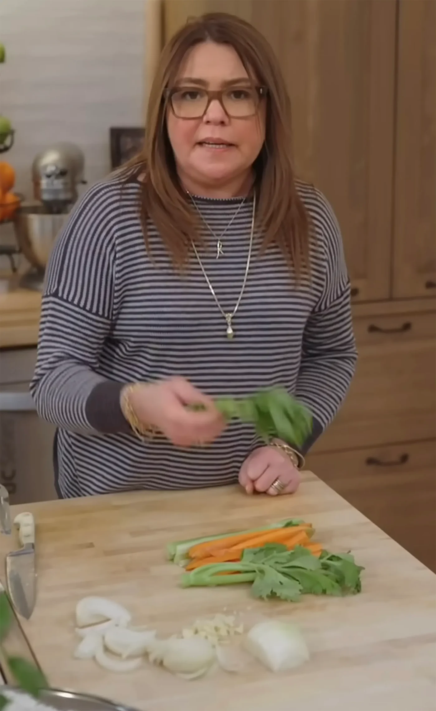 Rachael Ray in a kitchen, holding a bunch of herbs over a wooden cutting board with chopped vegetables.