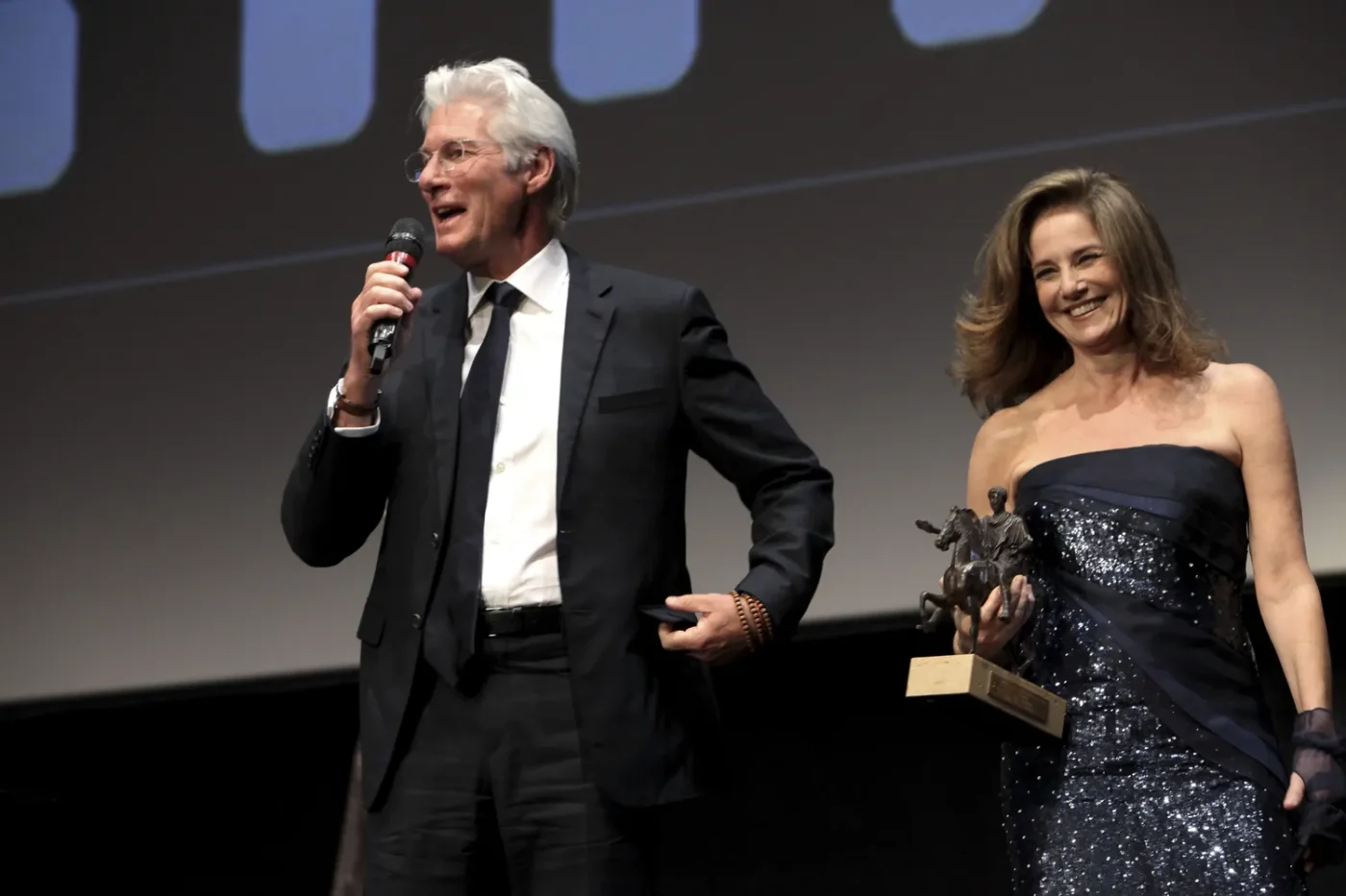 Richard Gere speaking into a microphone Debra Winger holding an award trophy on stage