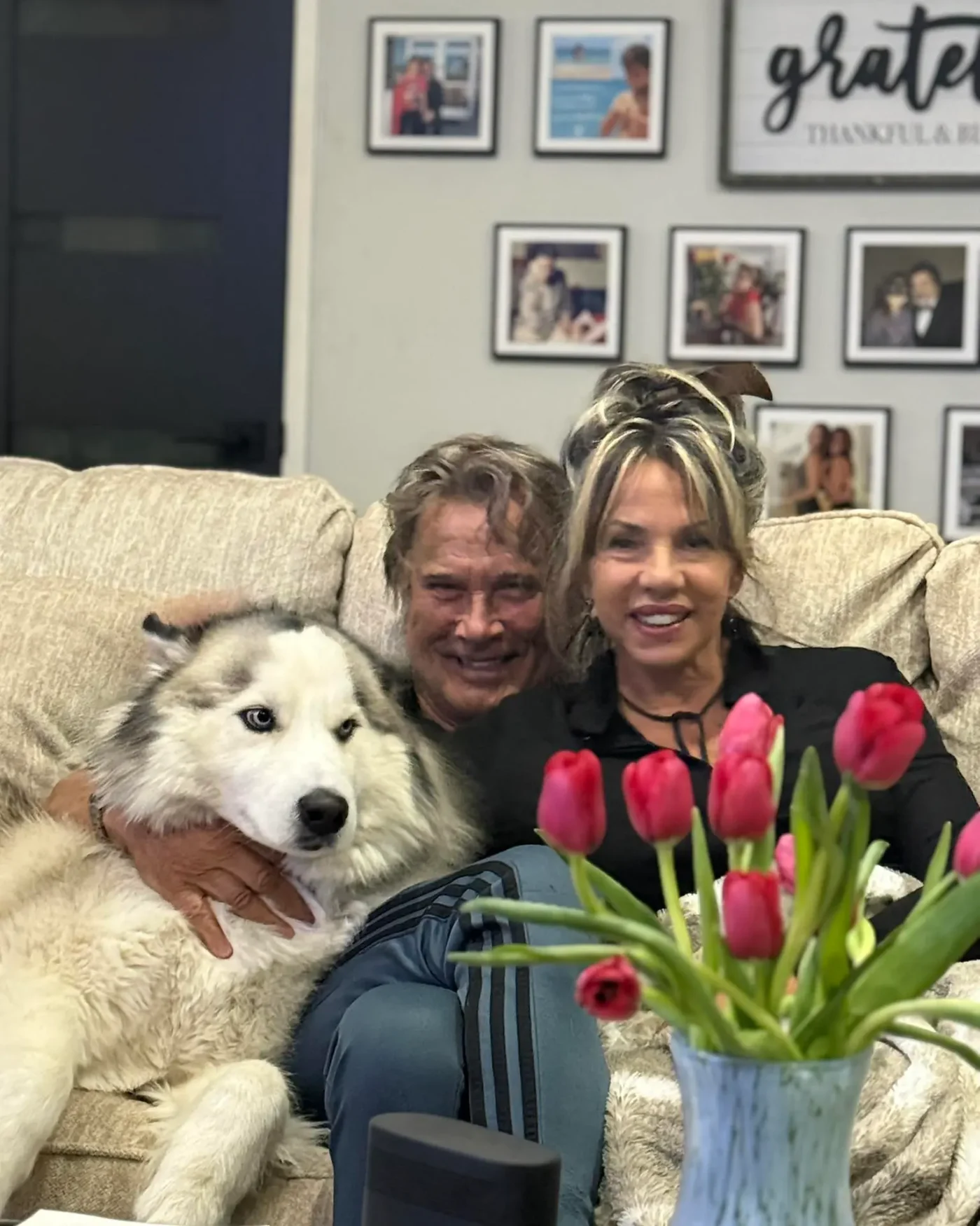 Richard Norton and his wife Judy sitting on a couch with a husky and a vase of red tulips.