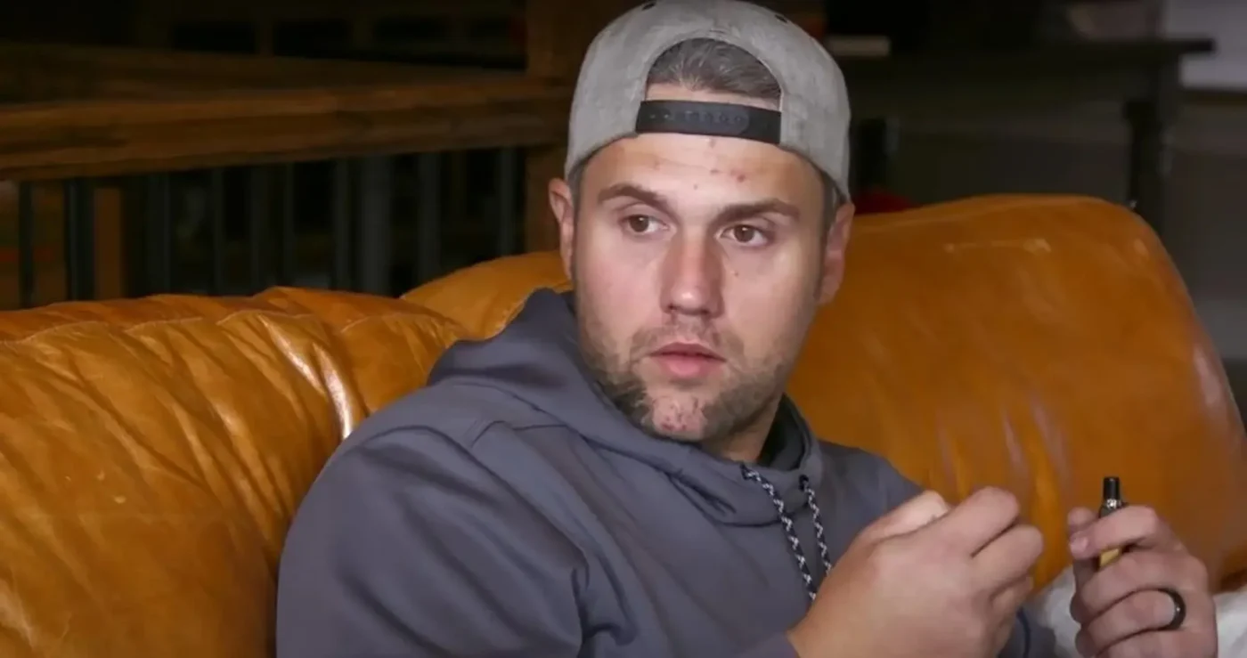 Ryan Edwards sitting on a leather couch, wearing a grey hoodie and baseball cap backward.