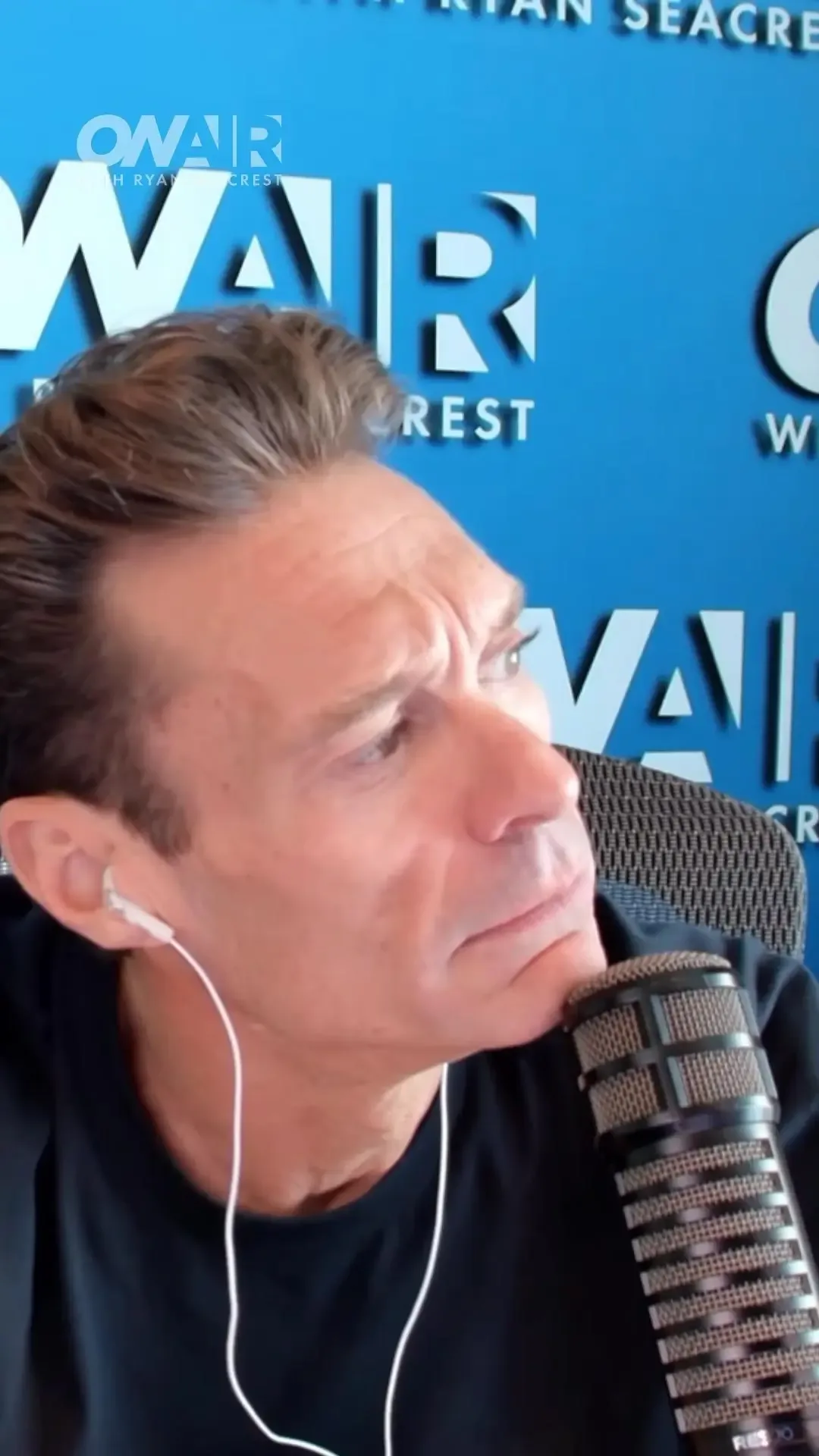 Ryan Seacrest sits in front of an On Air with Ryan Seacrest logo while speaking into a microphone.