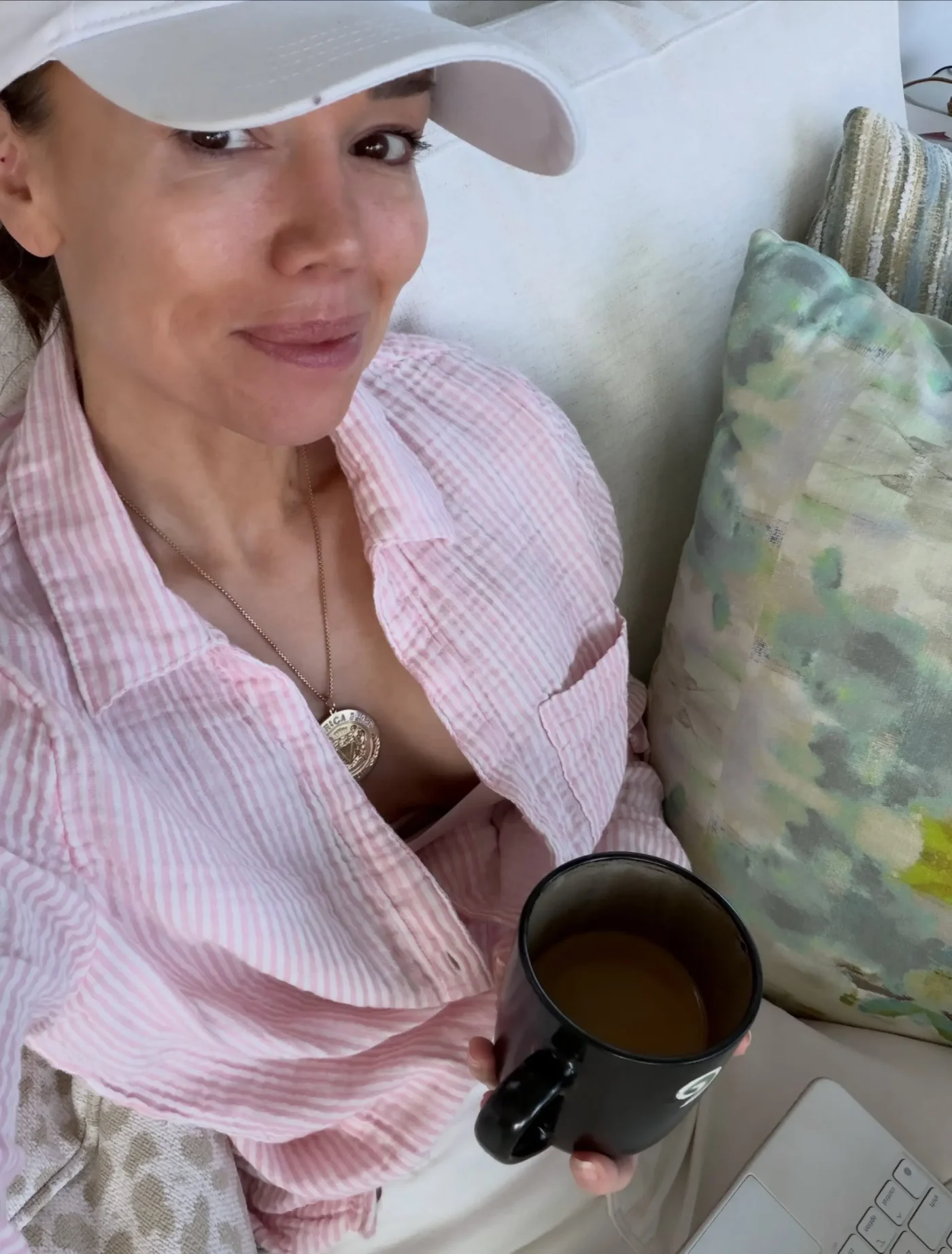 Sara Gore wearing a white cap and a pink-and-white striped shirt, holding a black mug of coffee on a couch.