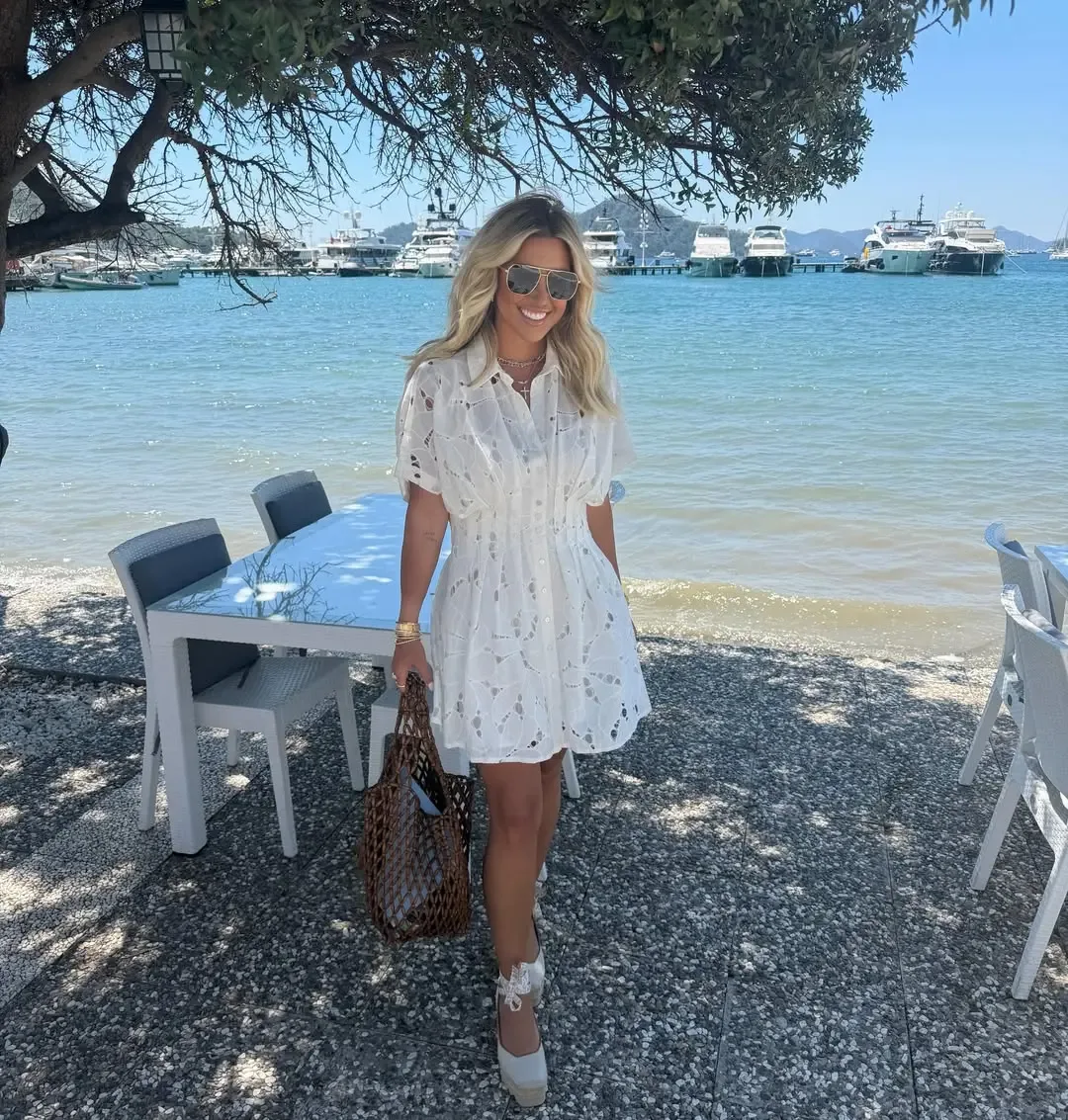 Savannah Chrisley stands by a waterfront restaurant holding a brown woven bag, wearing a white lace dress and sunglasses.