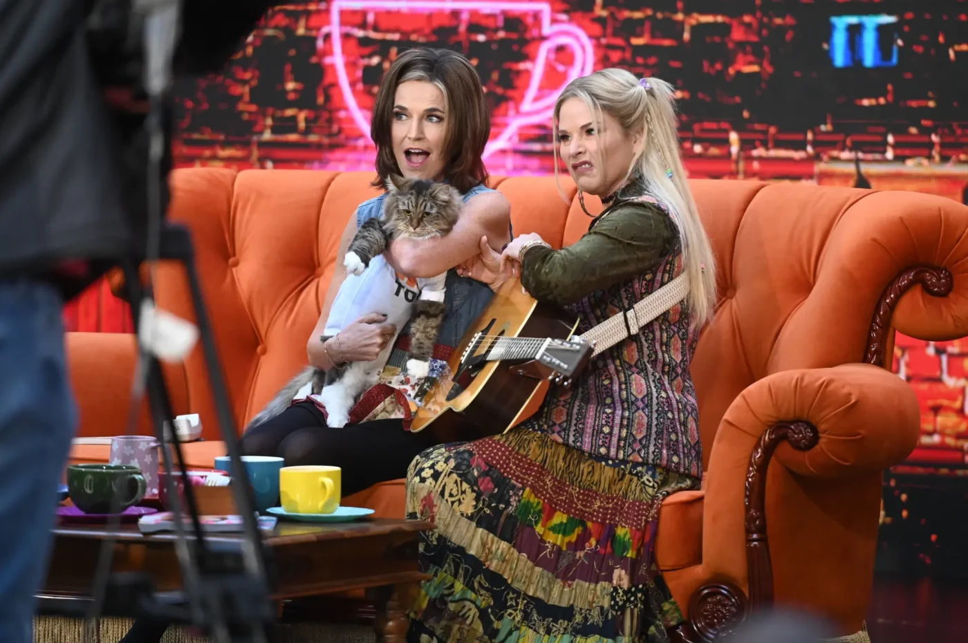 Savannah Guthrie and Jenna Bush Hager dressed as characters from the sitcom Friends on an orange sofa, with Guthrie holding a cat and Bush Hager holding a guitar.