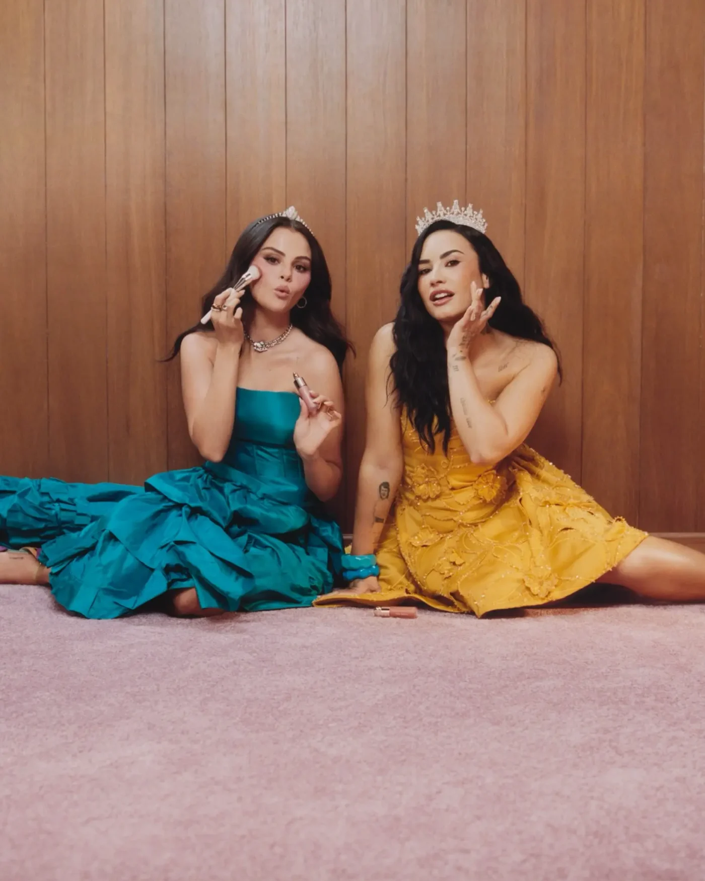 Selena Gomez and Demi Lovato wearing tiaras and dresses while posing with makeup.