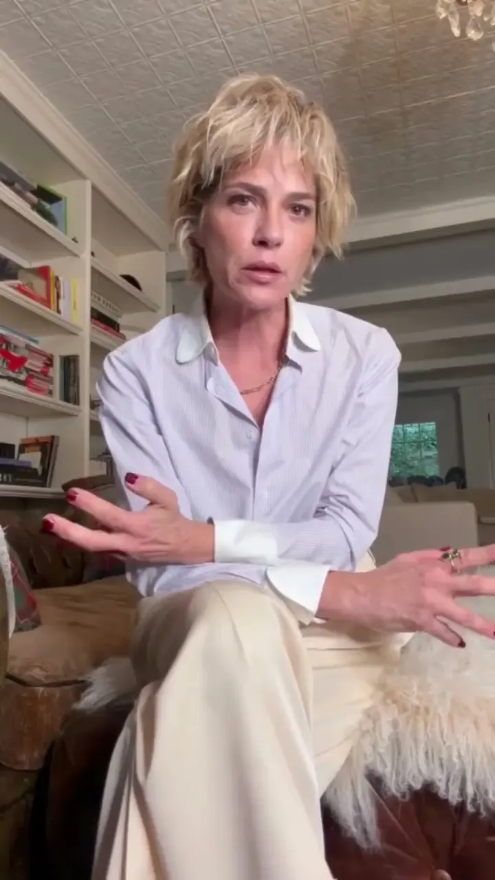 Selma Blair sits indoors, wearing a light-colored button-down shirt and trousers, gesturing with her hands while speaking.