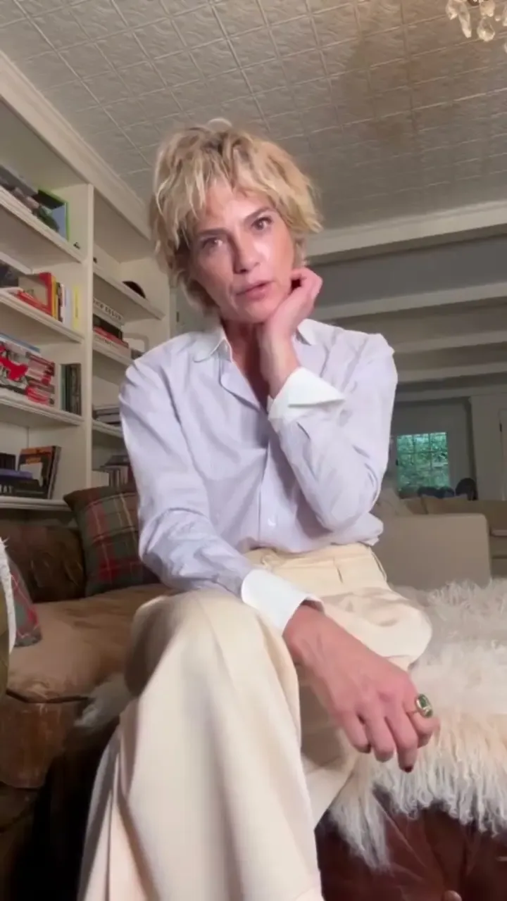 Selma Blair sitting indoors, wearing a white button-down shirt and light-colored pants.