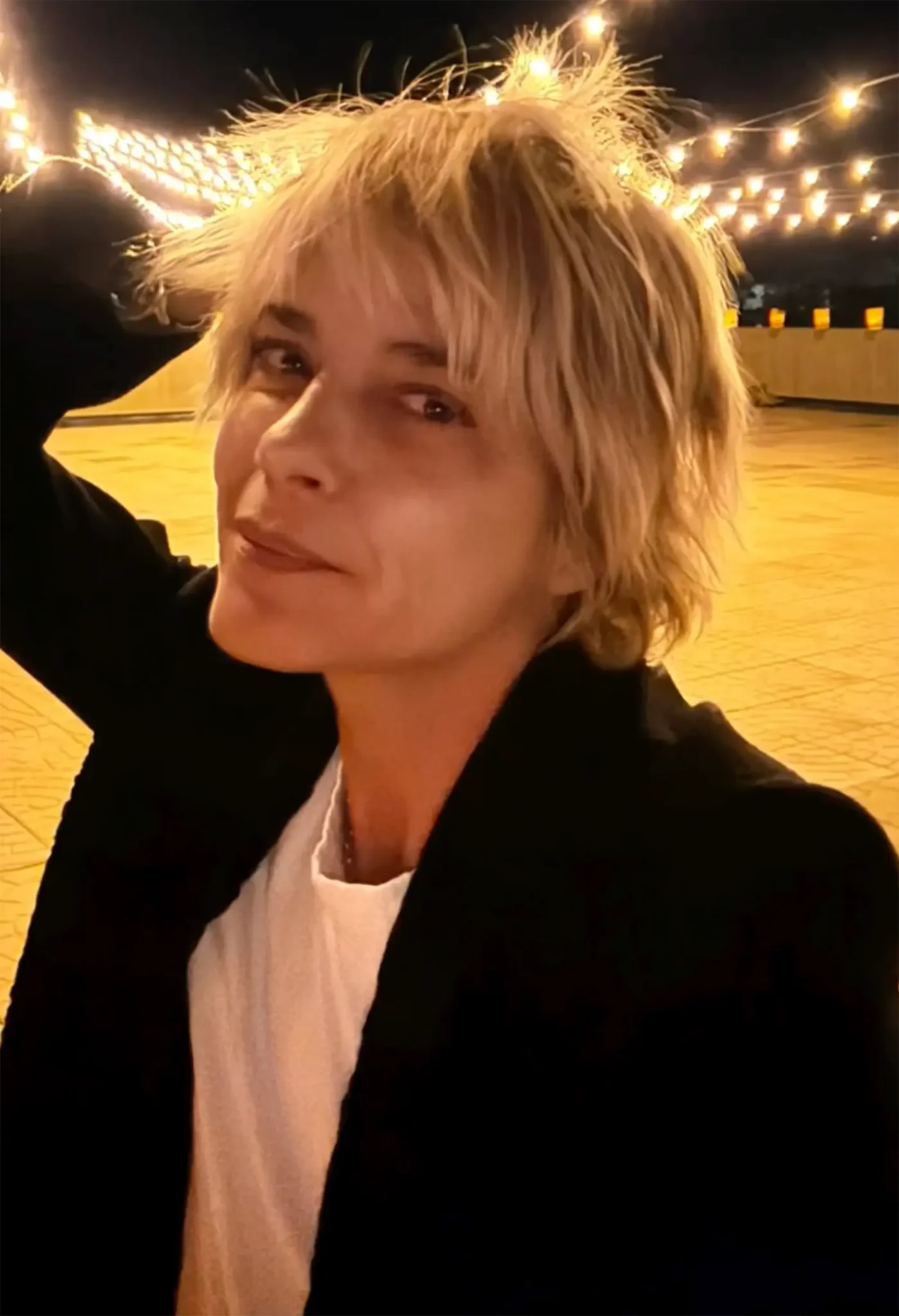 Selma Blair with short blonde hair and bangs, wearing a white t-shirt and black jacket, poses against a backdrop of outdoor string lights.