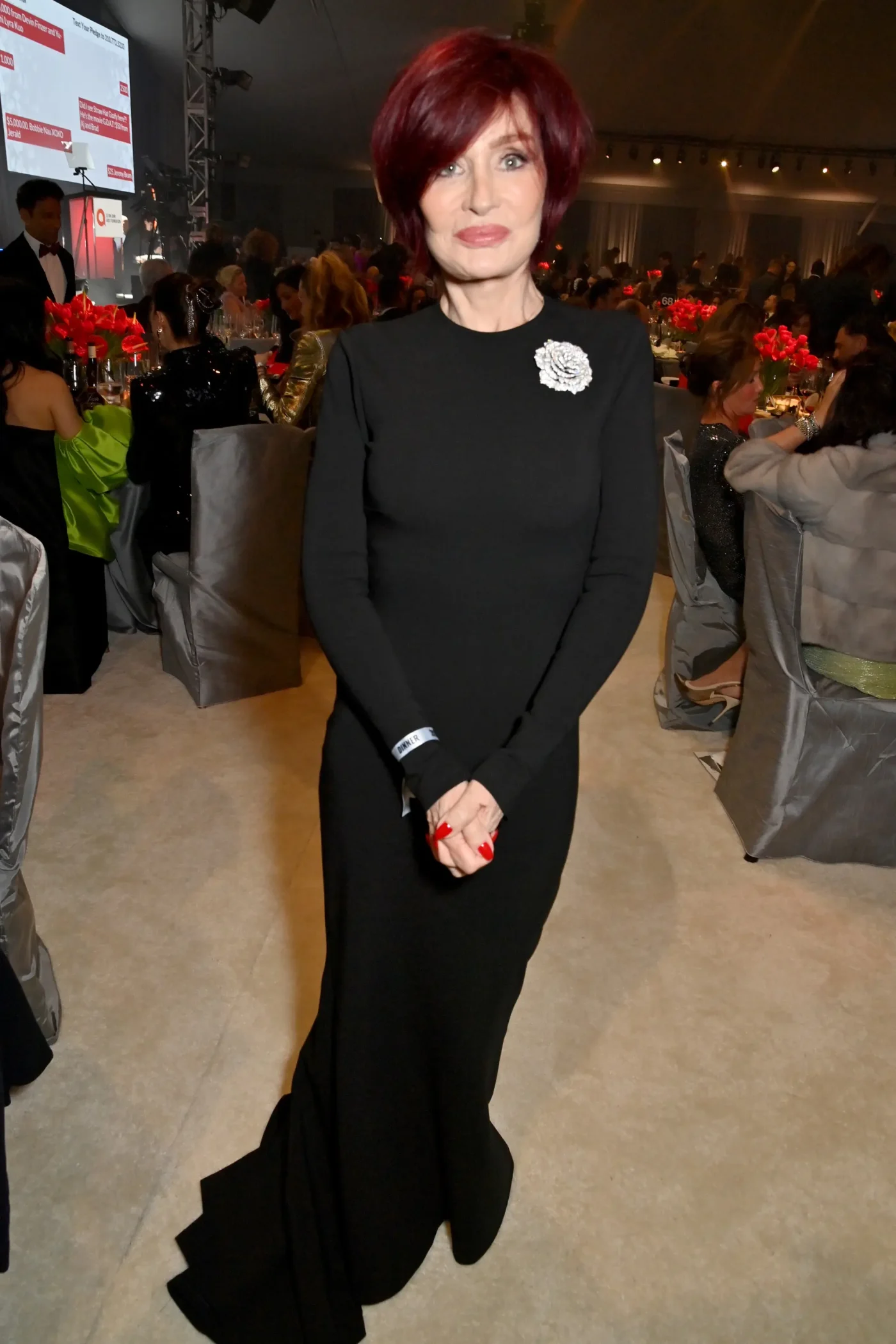 Sharon Osbourne stands at a gala, wearing a long-sleeved black dress with a floral brooch, her vibrant red hair styled short.