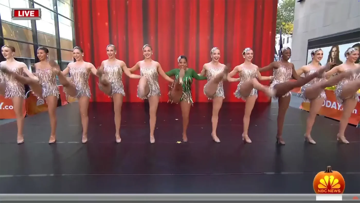 Sheinelle Jones and a line of dancers perform a high-kick routine in front of a red curtain.