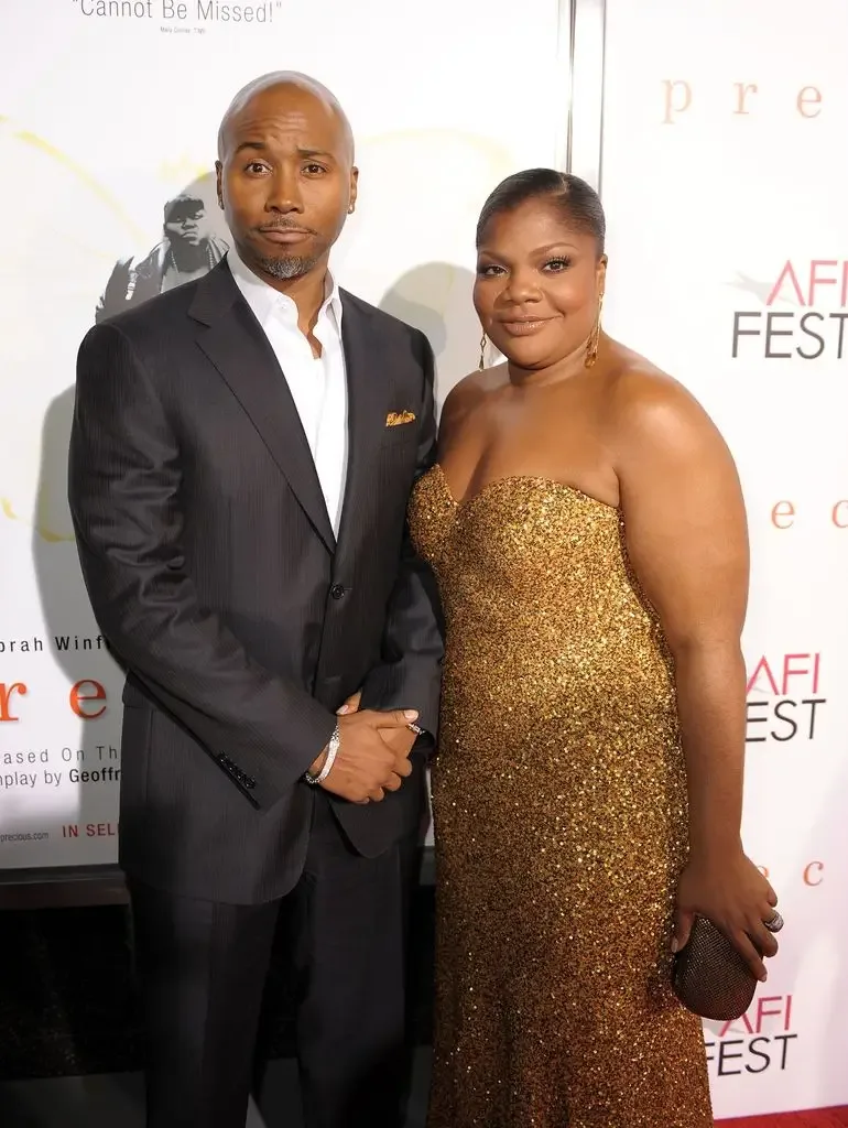 Sidney Hicks in a dark suit stands beside Mo'Nique, who wears a strapless gold sequined gown, at a red carpet event.
