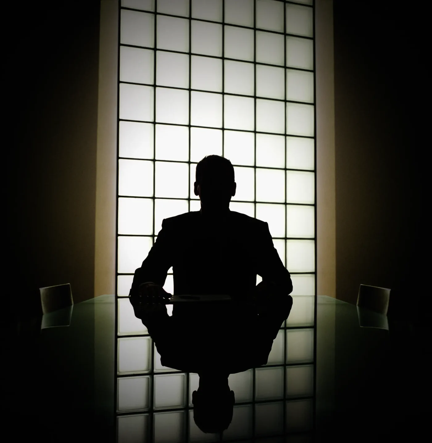 Silhouette of a businessman seated at a conference table, backlit by a grid window.