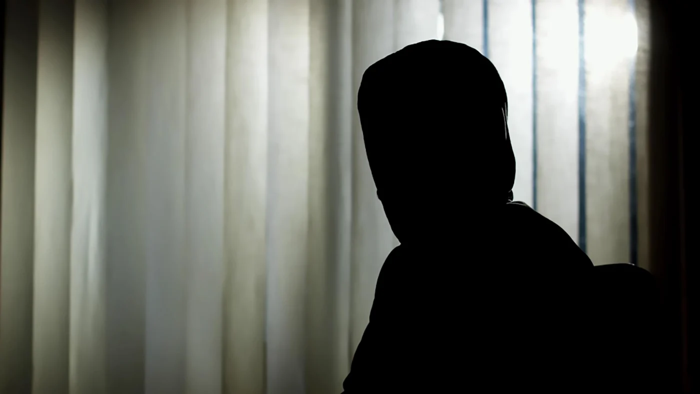 Silhouette of a witness in a protective disguise against a lit window with vertical blinds.