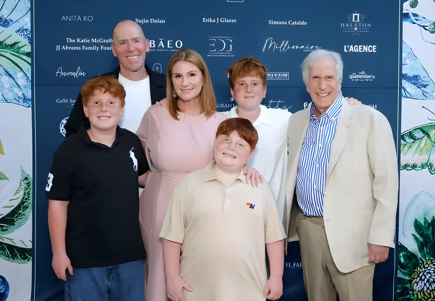 Six smiling people, four of them red-haired boys and men and a woman in a pink dress, pose close together in front of a blue sponsor backdrop, with Henry Winkler in a cream blazer and striped shirt on the right.