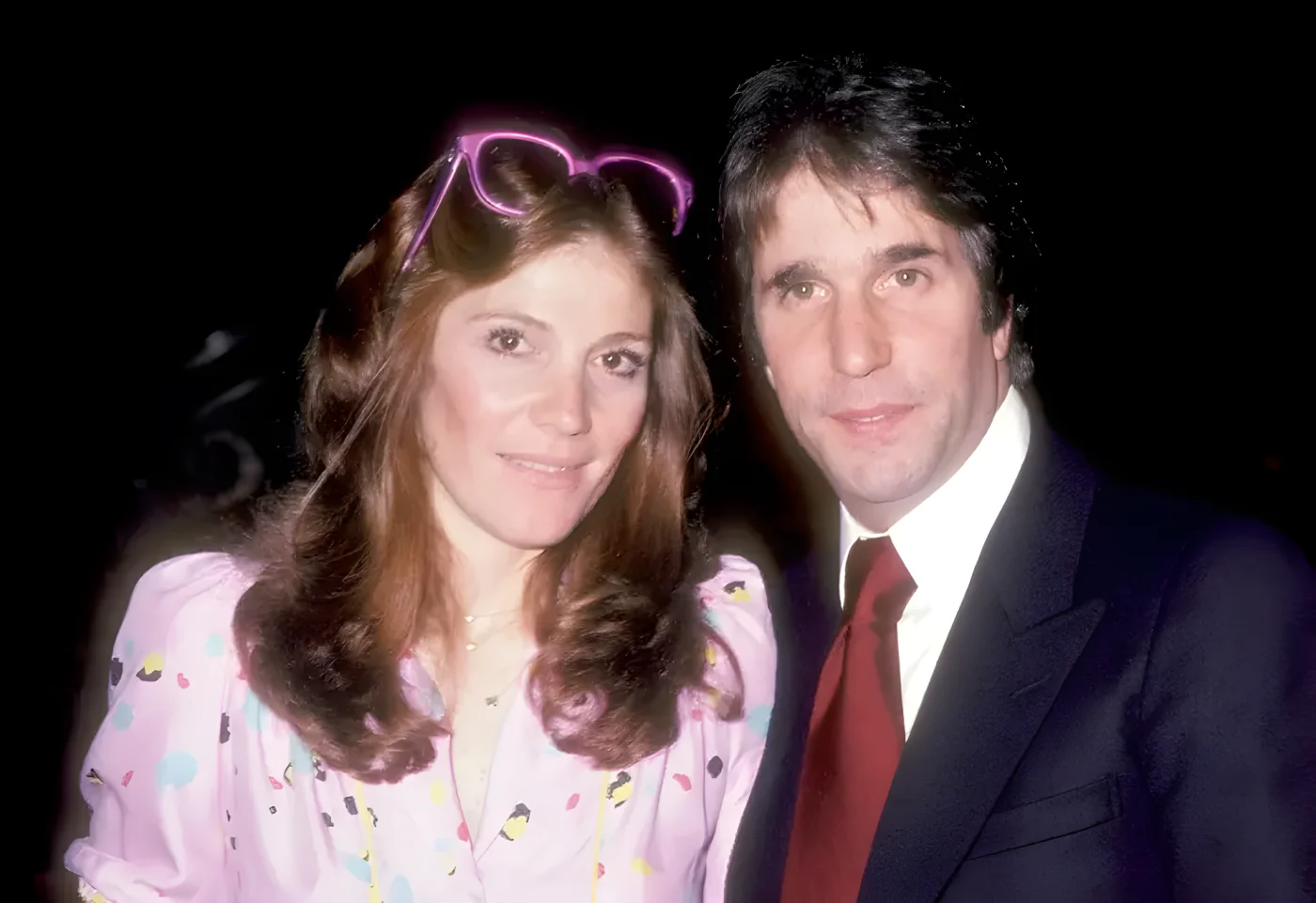 Stacey Weitzman, with auburn hair and pink sunglasses on her head, poses cheek to cheek with Henry Winkler, who wears a dark suit and red tie, against a dark background.