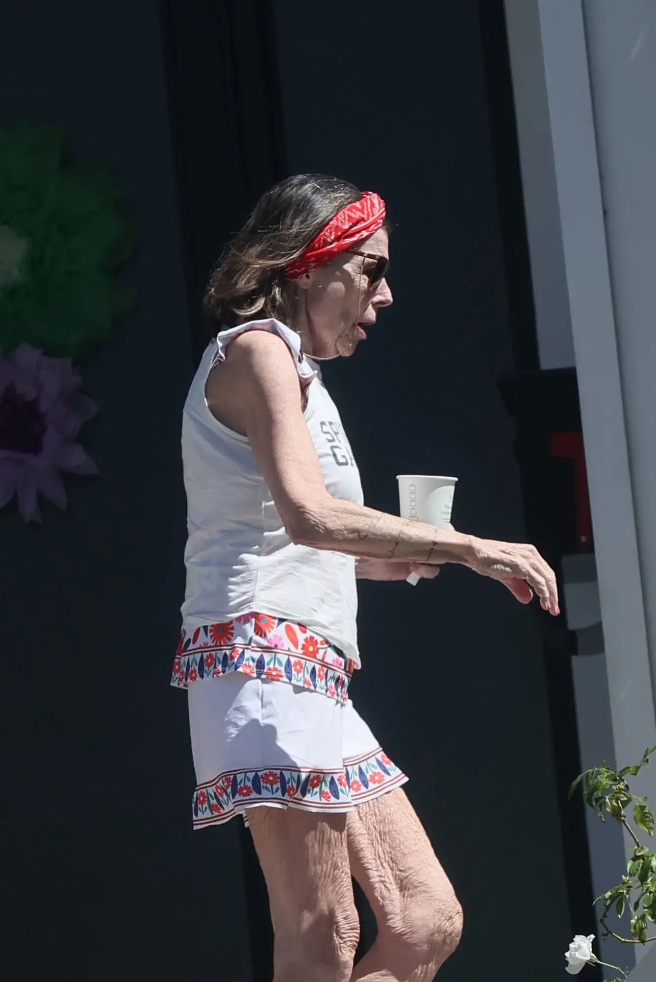 Susan St. James wearing a white outfit and red headband, holding a paper cup outside her home.