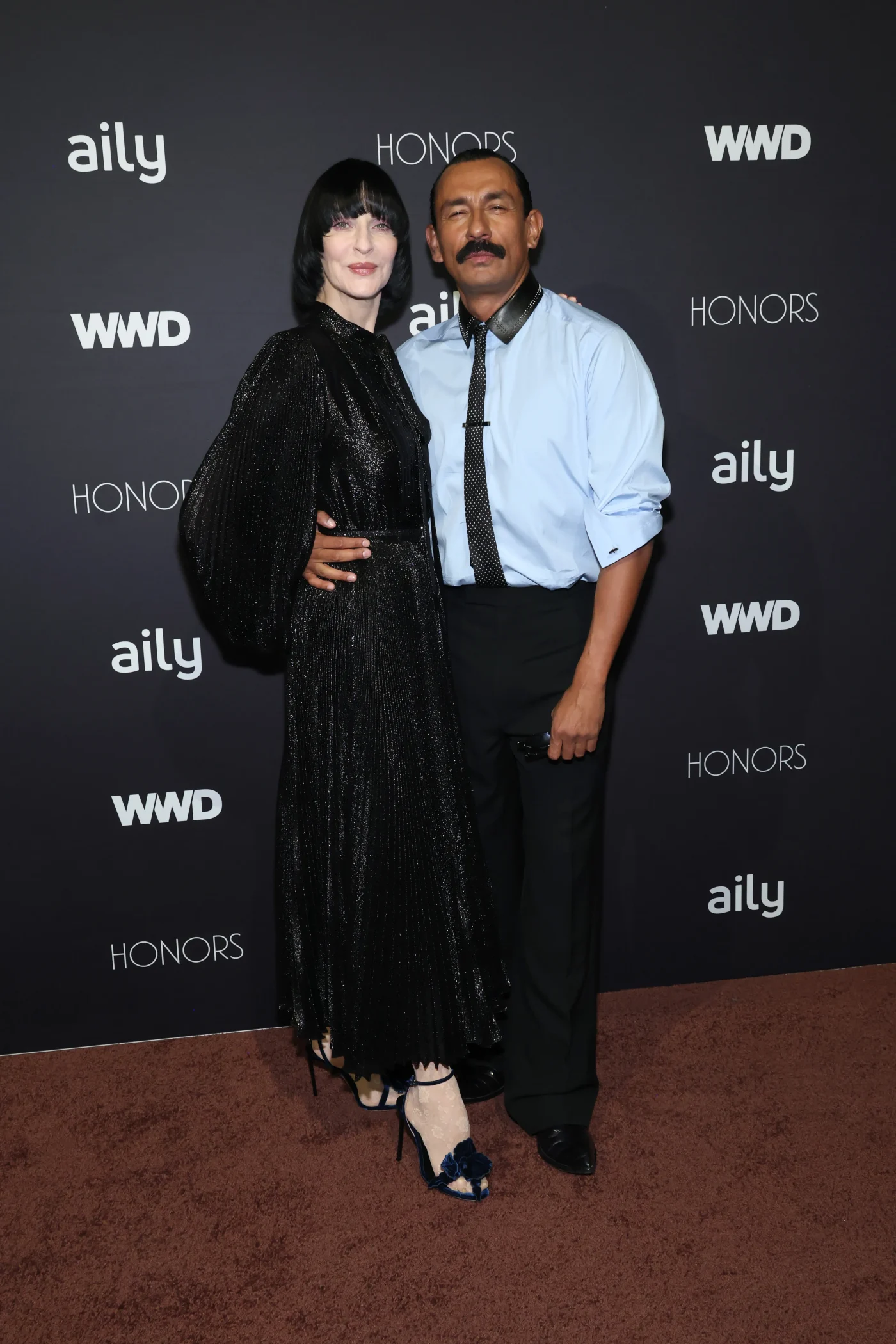 Susie Cave and Haider Ackermann posing in front of a WWD Honors step-and-repeat backdrop.