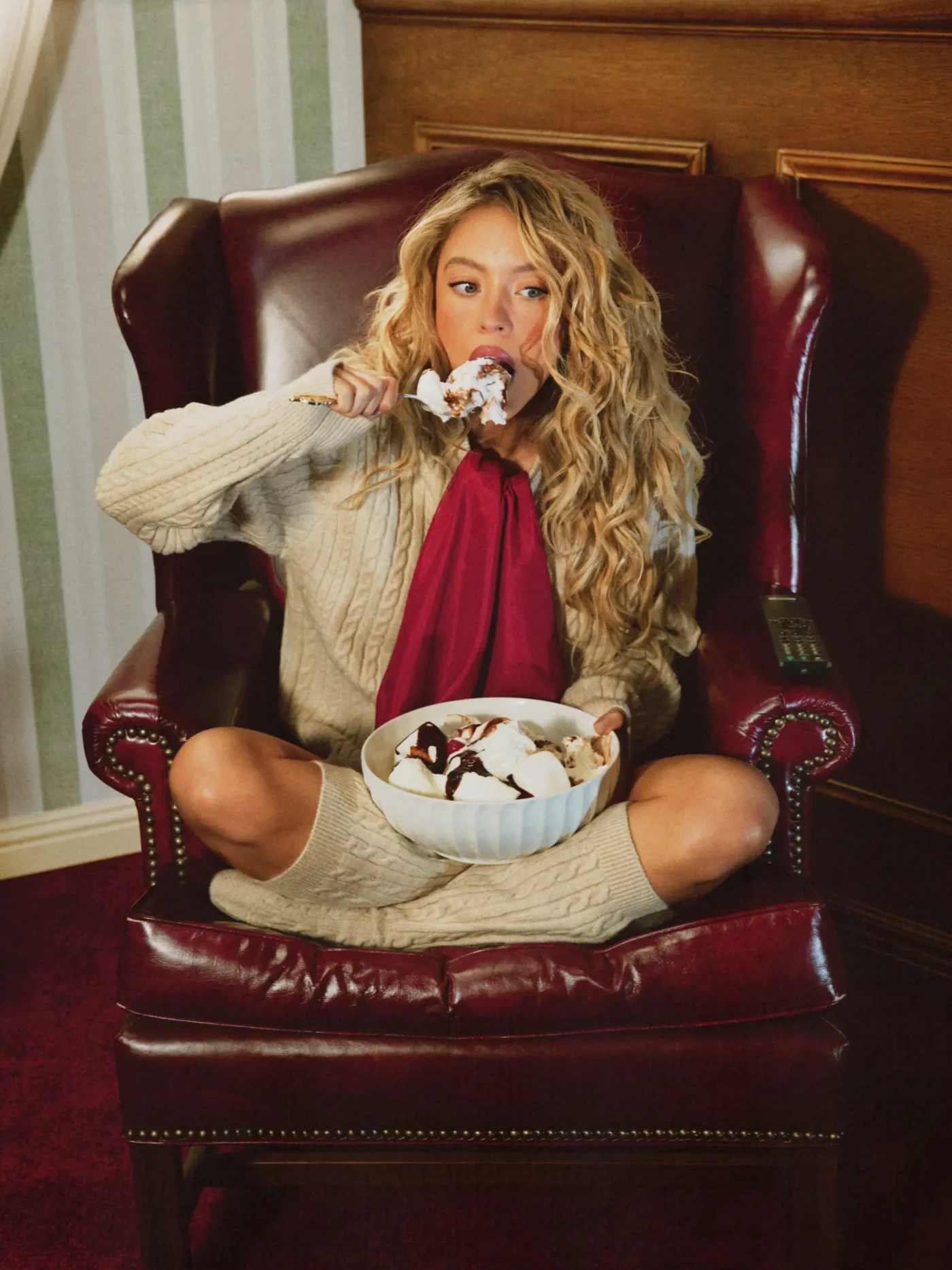 Sydney Sweeney sitting cross-legged in a red leather armchair, eating ice cream from a large bowl.
