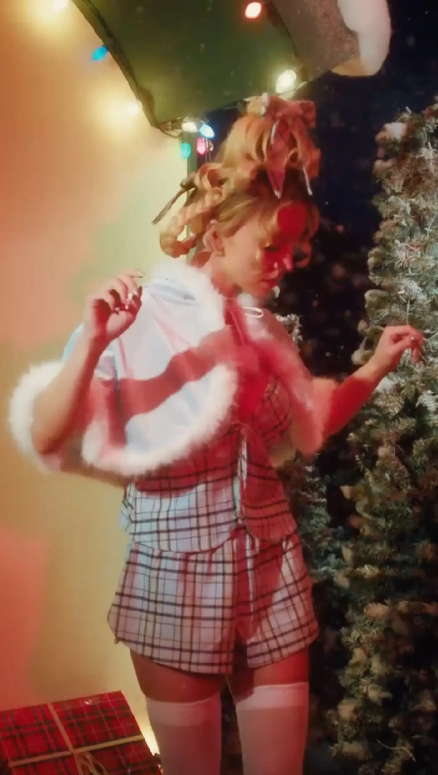 Sydney Sweeney with an updo hairstyle wearing a plaid outfit and a capelet with white trim near a Christmas tree.