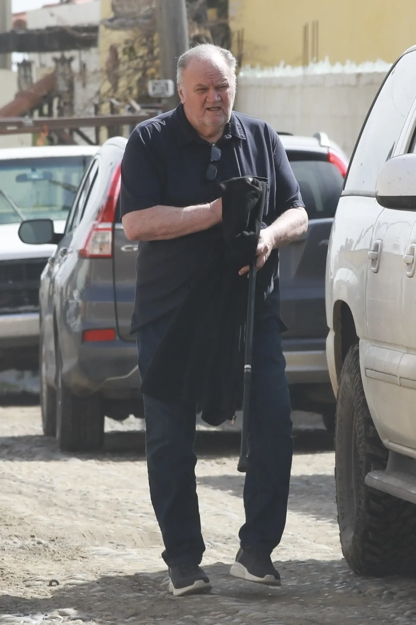 Thomas Markle walks down a cobbled street in Mexico, holding a jacket and a cane between parked cars.