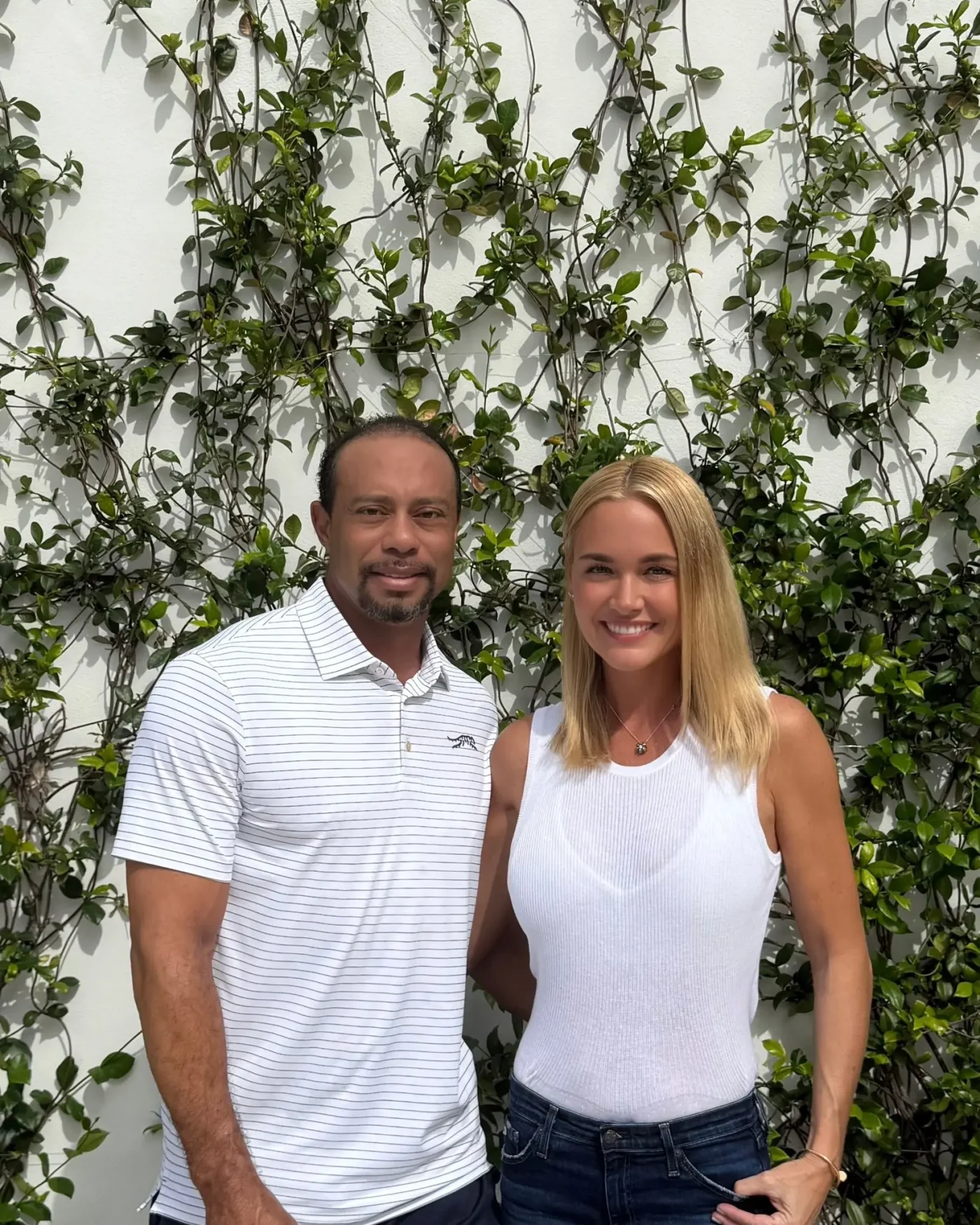 Tiger Woods and Vanessa Trump stand together smiling in front of a white wall covered in climbing green vines.