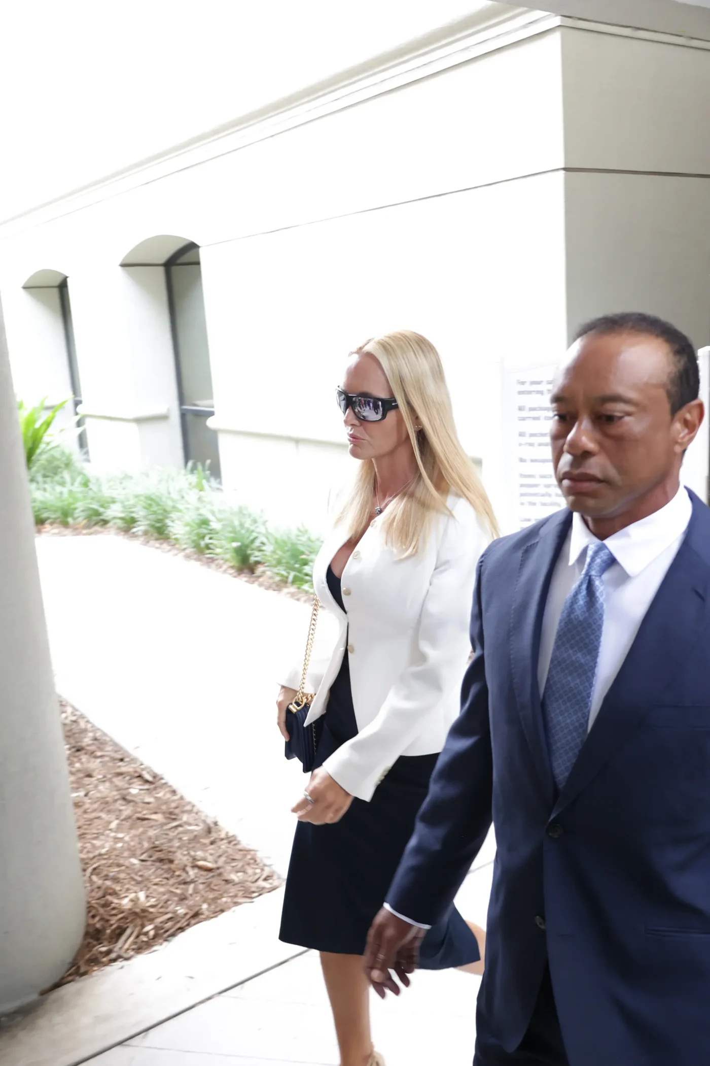 Tiger Woods and Vanessa Trump walking outdoors, with Woods in a suit and Trump in a white jacket and sunglasses.