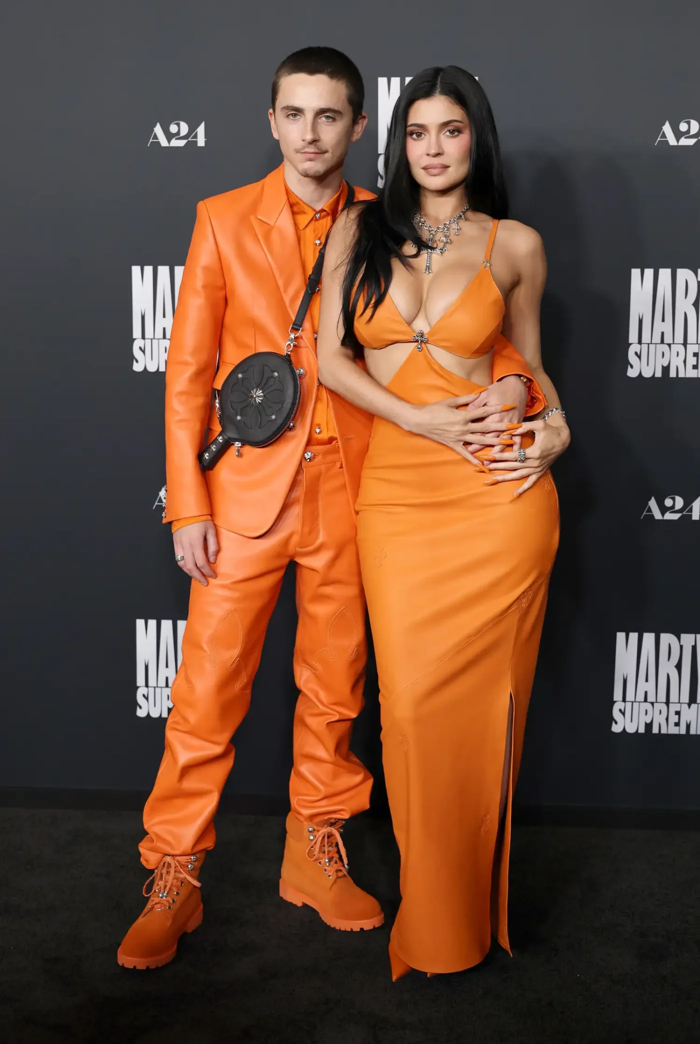 Timothée Chalamet and Kylie Jenner pose for a photo on the red carpet. Chalamet is dressed in an all-orange leather suit with matching work boots and a black cross-body bag. Jenner is dressed in a vibrant orange gown with large waist cutouts and a plunging neckline.