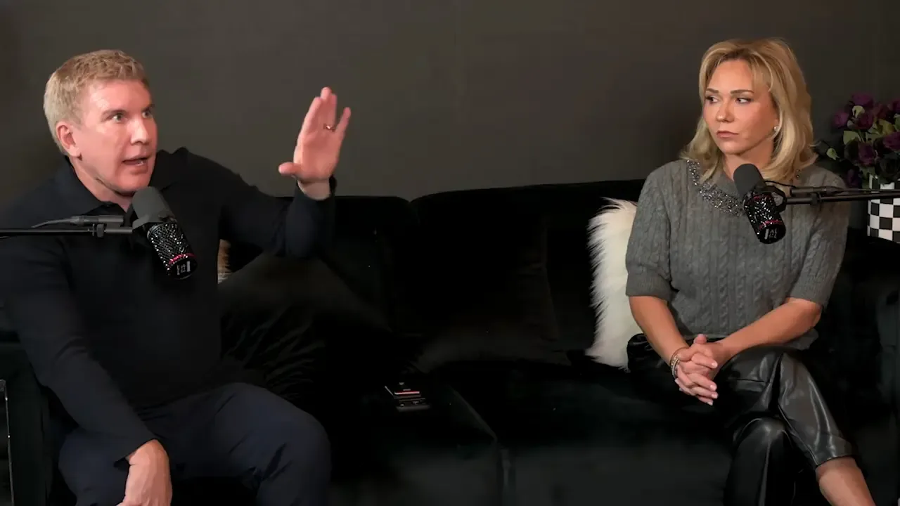 Todd Chrisley speaking with his hand raised, sitting on a couch next to a woman.