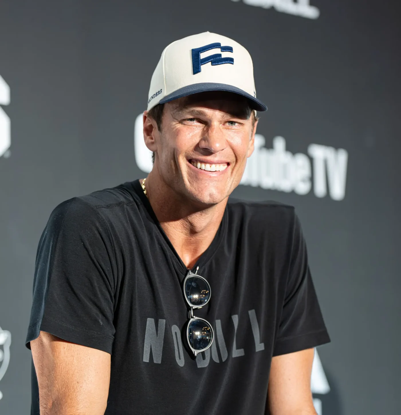 Tom Brady smiling while wearing a baseball cap and a black t-shirt.