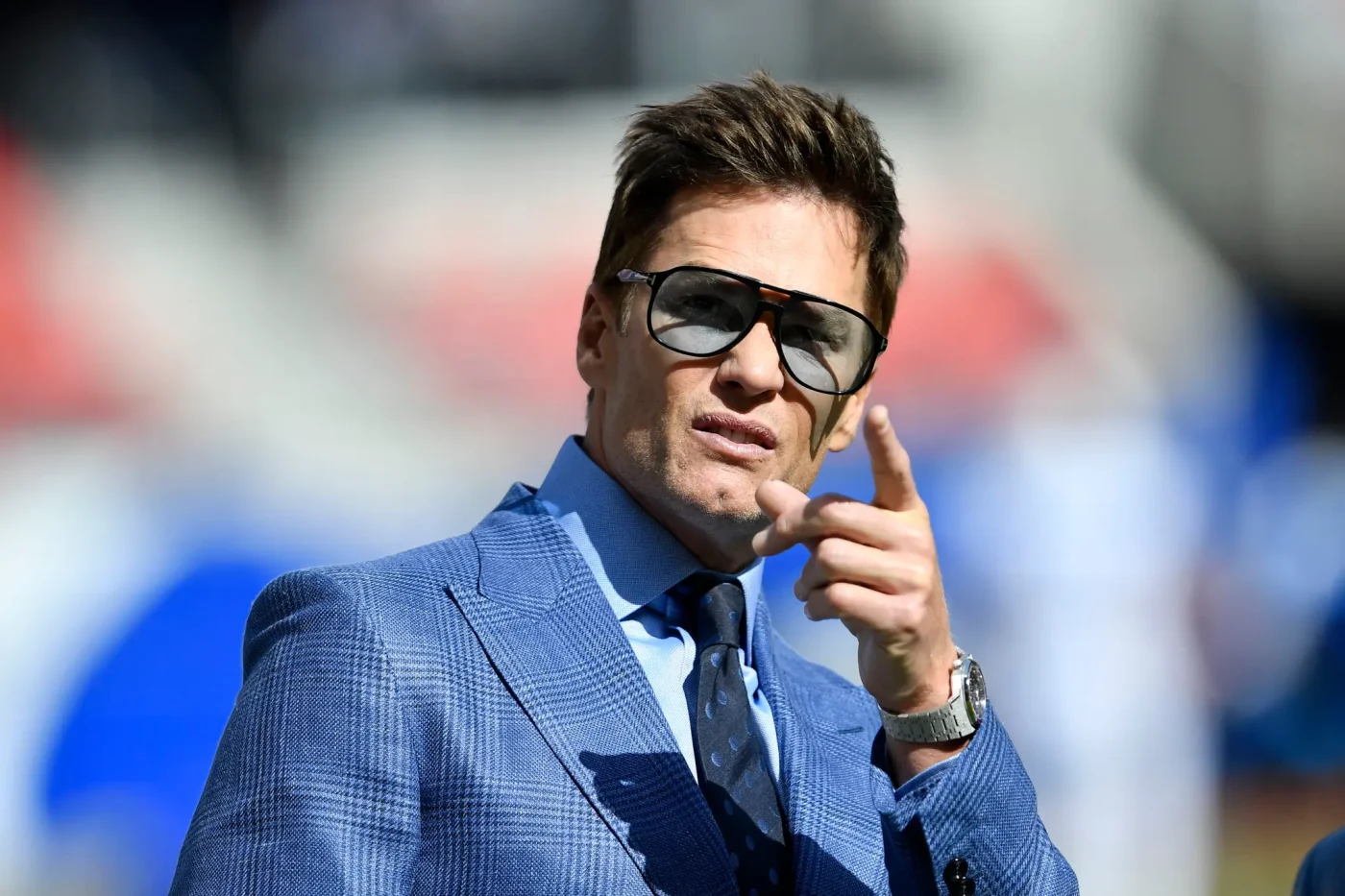 Tom Brady stands on the field wearing a blue suit and sunglasses, pointing forward.