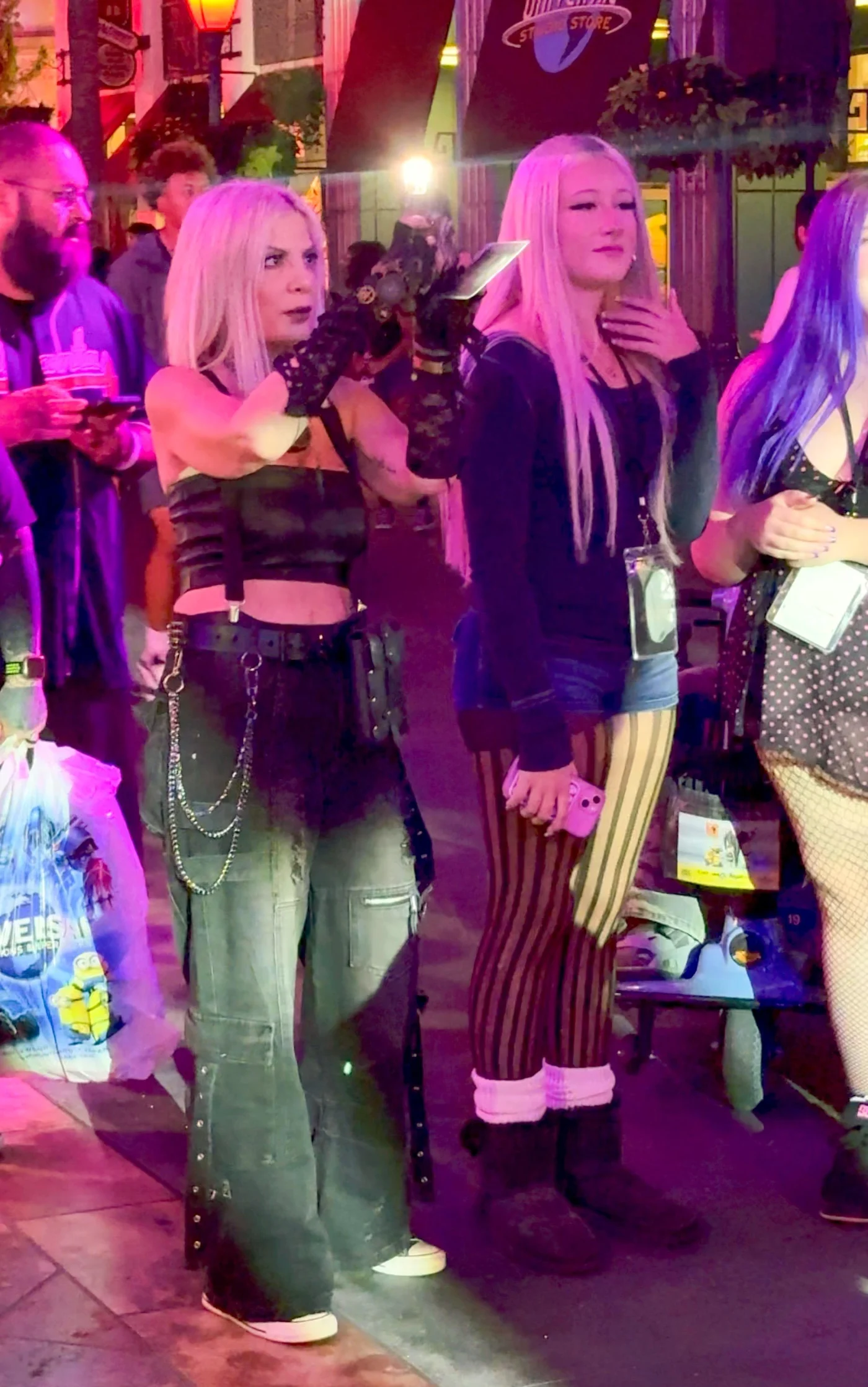 Tori Spelling with blonde hair, wearing a black tube top and cargo pants, poses outside at night.