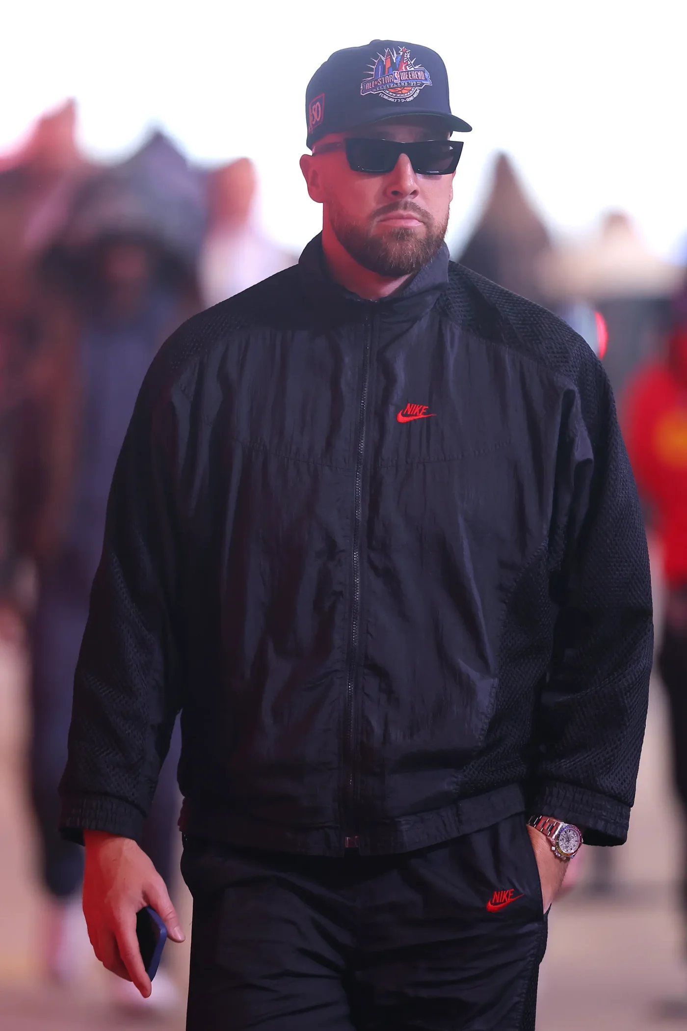 Travis Kelce arrives wearing a black Nike jacket, cap, and sunglasses.