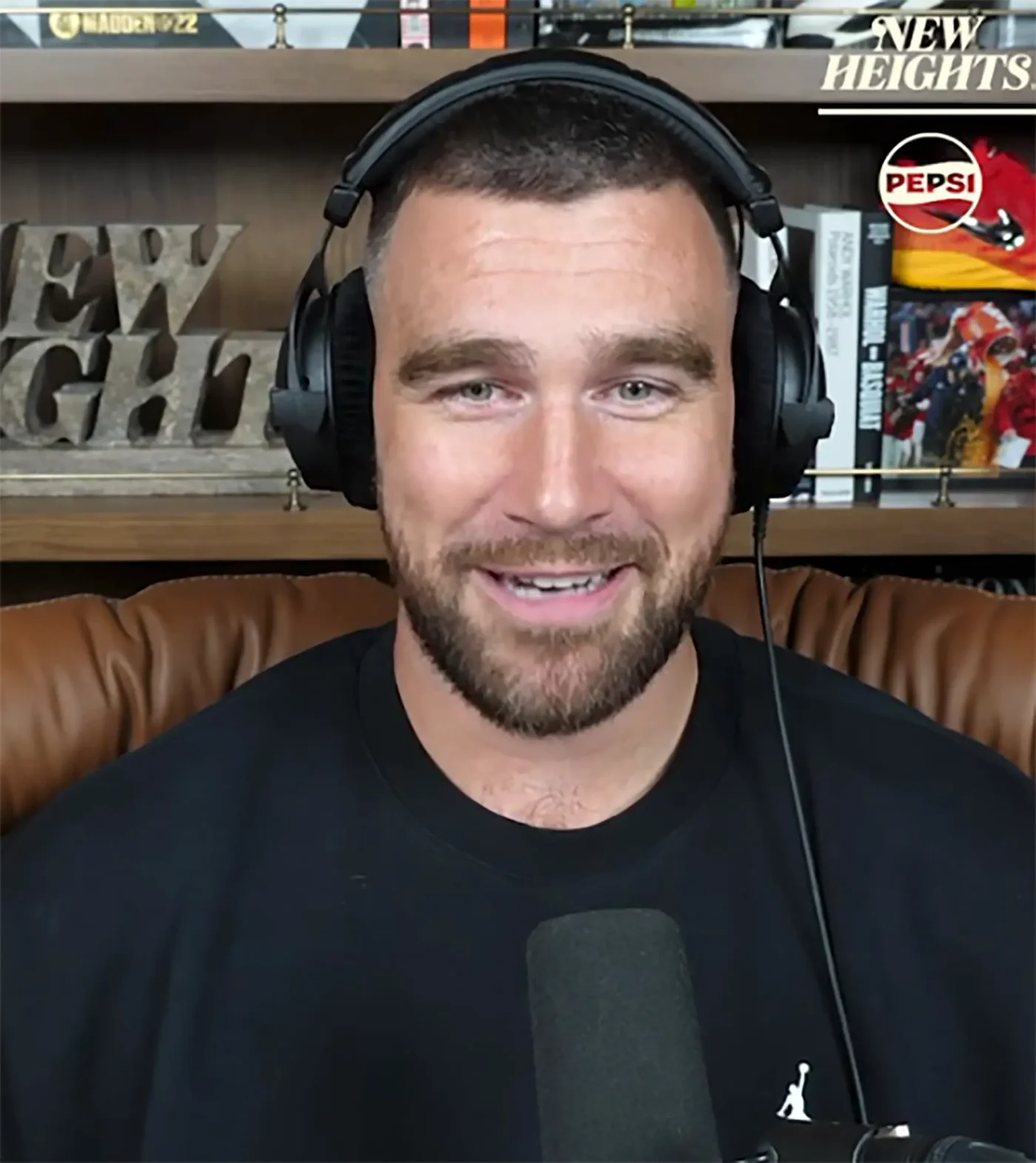 Travis Kelce smiling while wearing headphones and a black shirt in a recording studio.