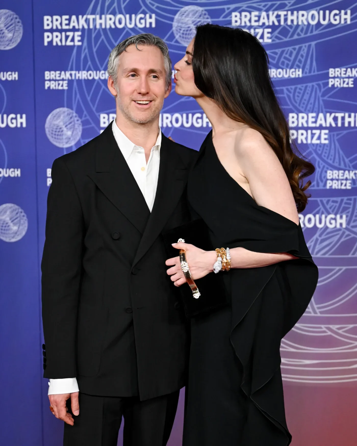 Two individuals on a red carpet at a breakthrough prize event one dressed formally receiving a kiss on the cheek