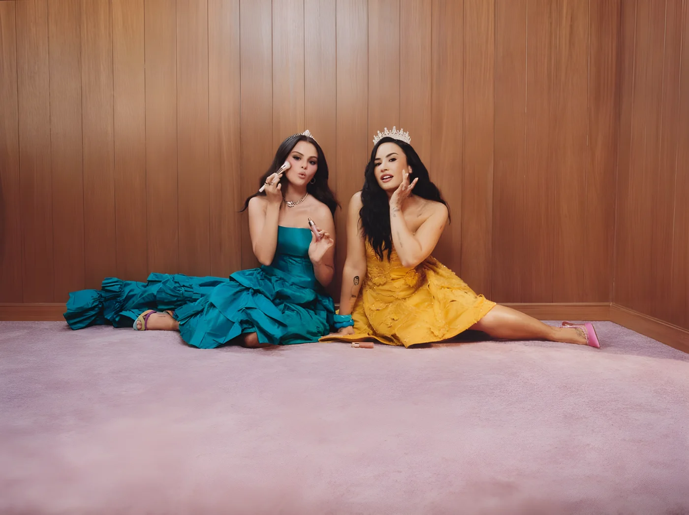 Two women dressed in colorful evening gowns apply makeup while seated on a plush surface in a cozy setting.