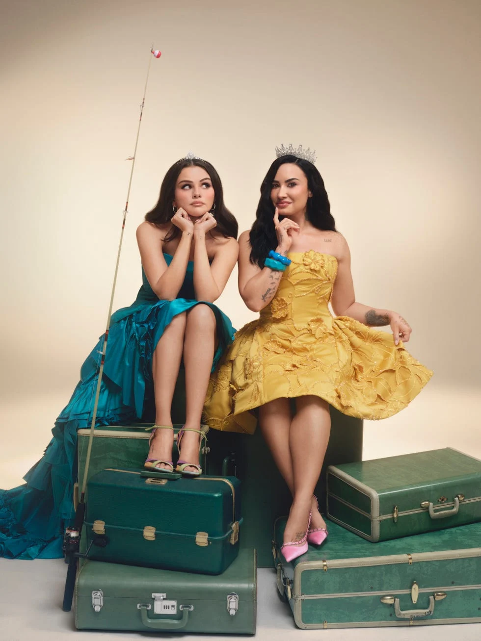 Two women in colorful gowns and tiaras sitting on green suitcases with playful expressions.