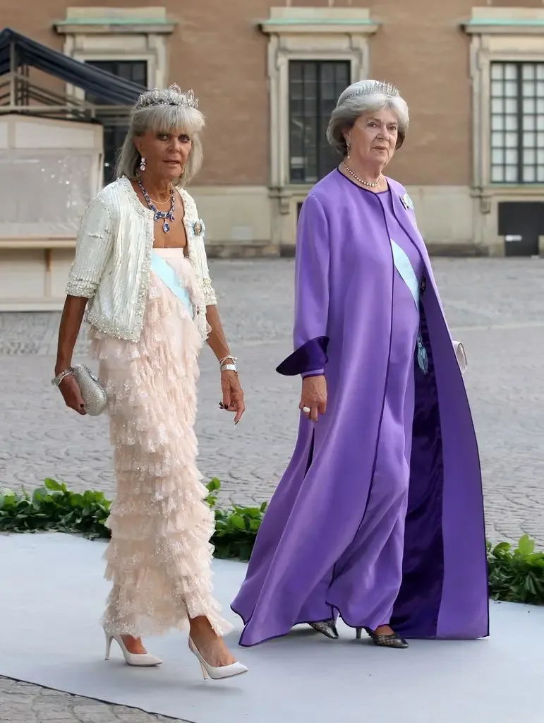 Two women in formal attire, one in a white dress and the other in a purple gown, walking on a path.