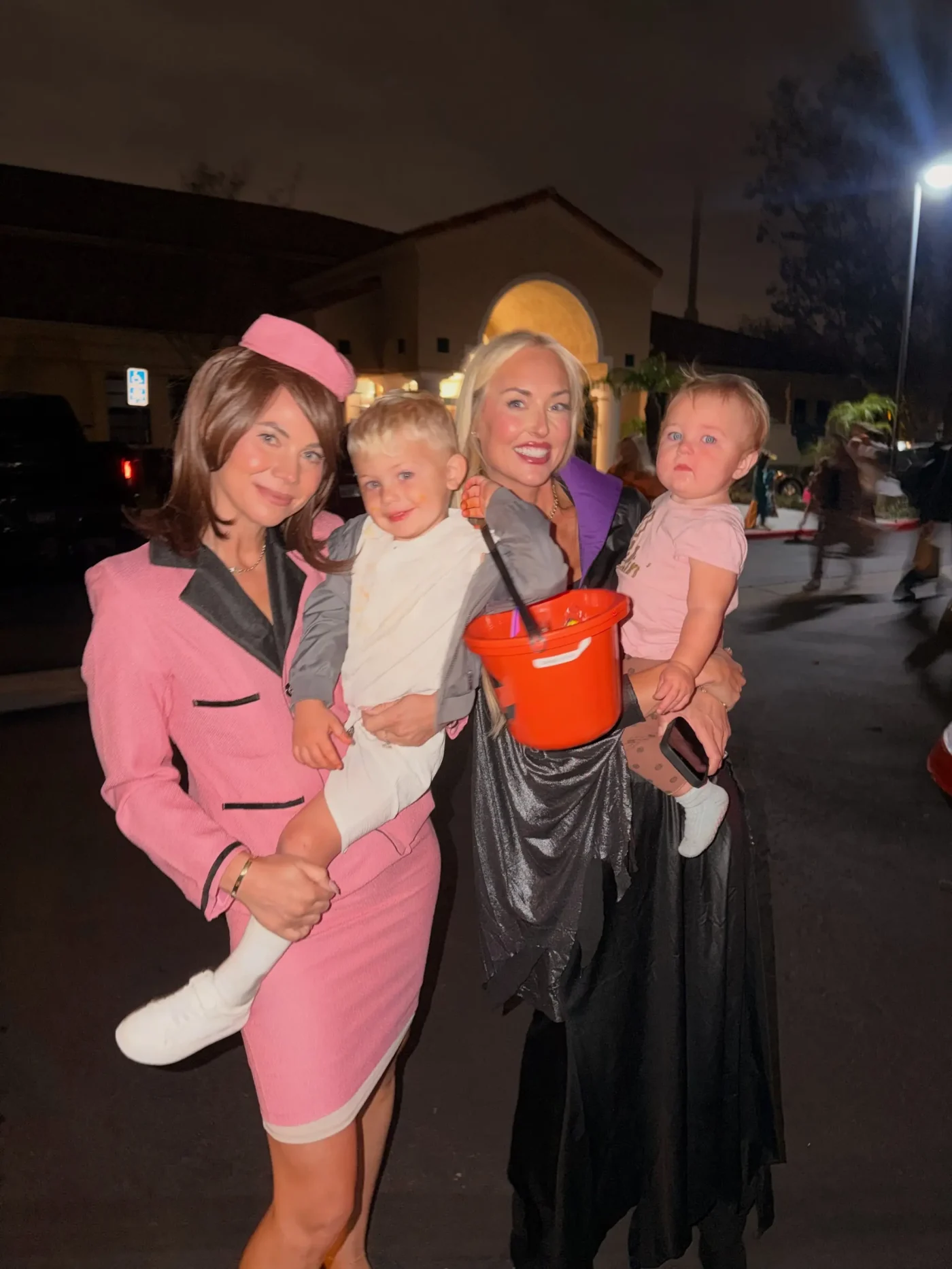 Two women posing in costumes outdoors at night while holding two toddlers, with one of the women holding an orange bucket.