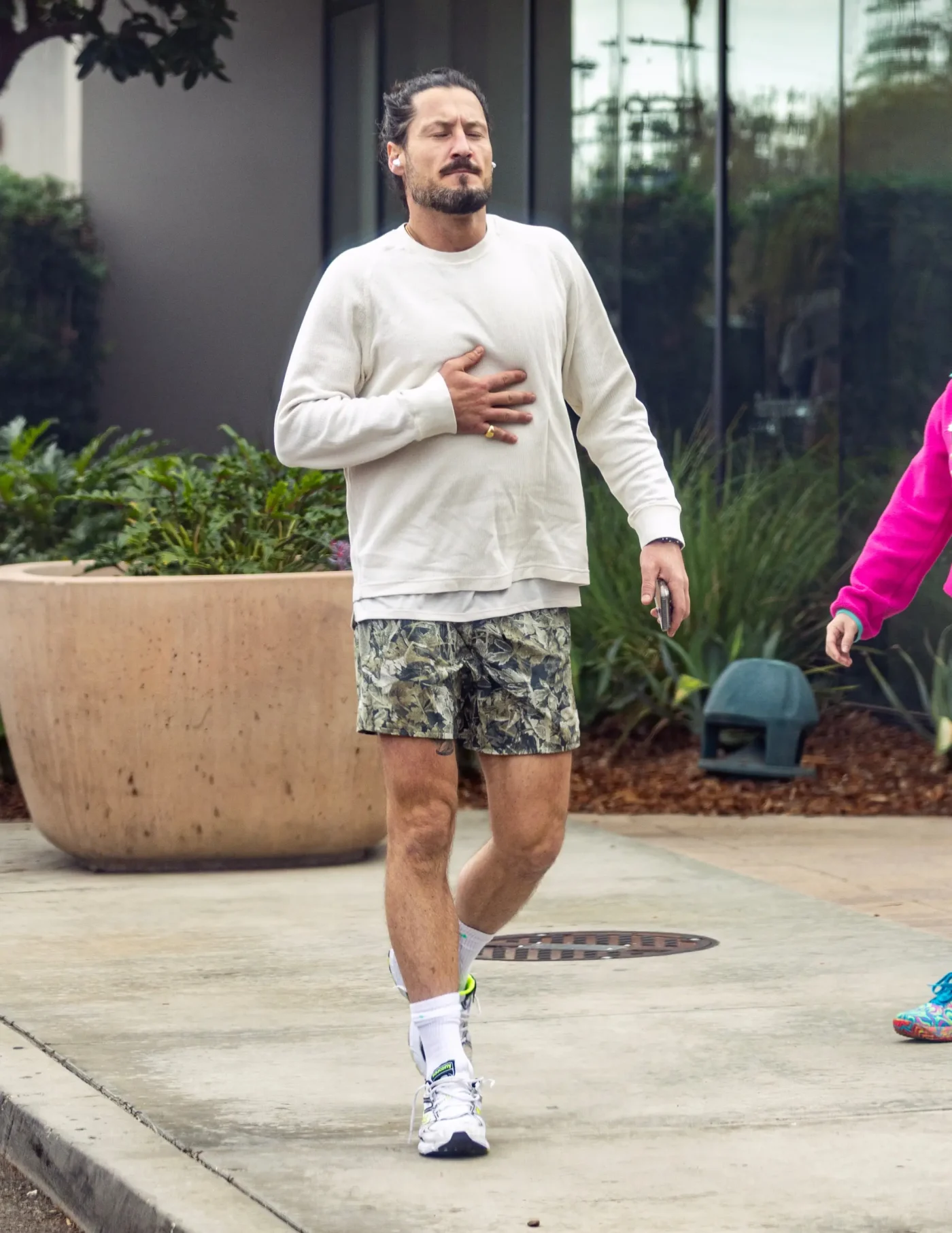 Val Chmerkovskiy walks outdoors, wearing a white long-sleeve shirt and patterned shorts, with his hand on his chest.