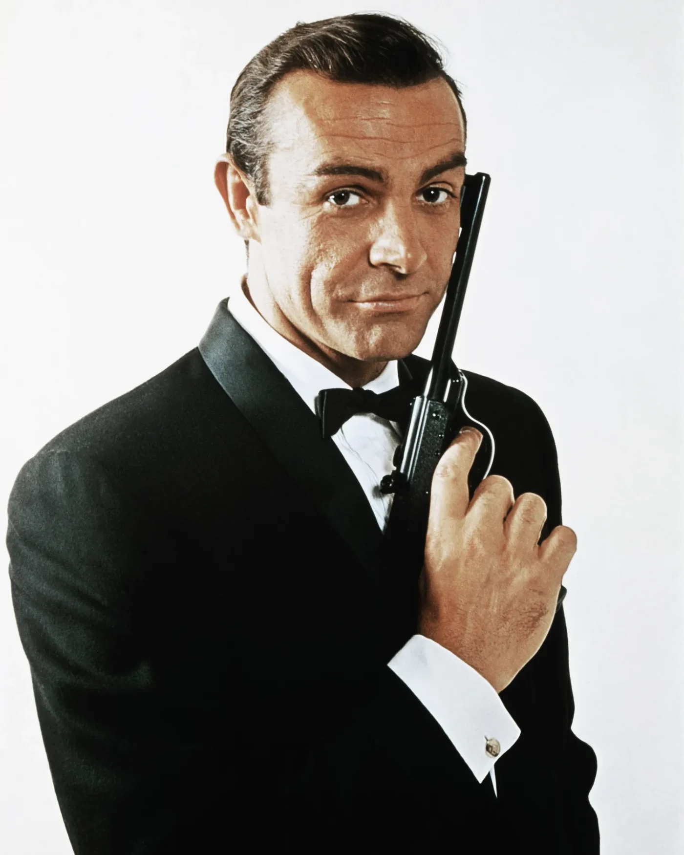 Waist-up portrait of Sean Connery, as James Bond, caressing the barrel of a gun against the side of his face. Connery is wearing a tuxedo and bow tie and smiling slightly.