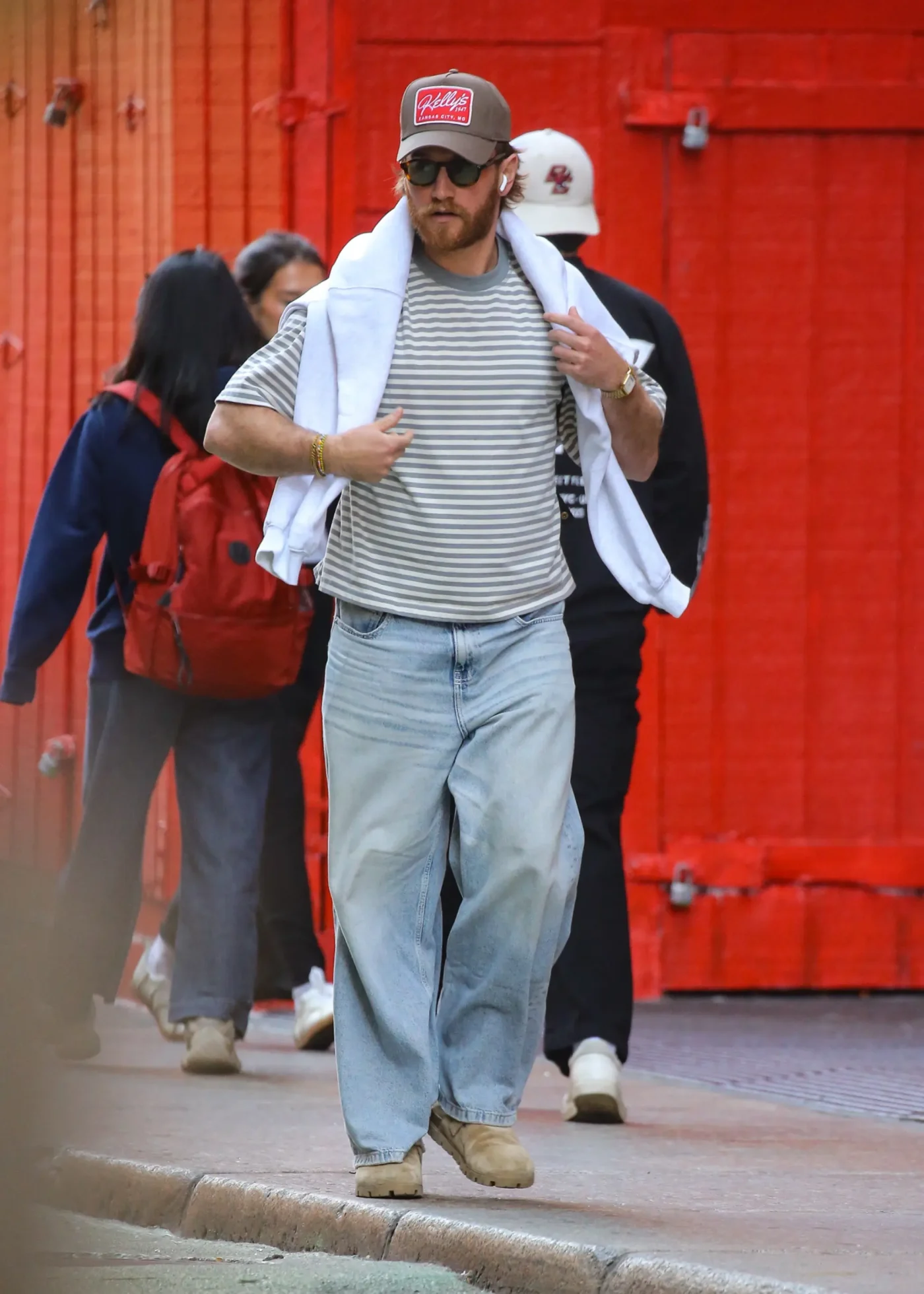 West Wilson is spotted walking in New York City wearing a striped t-shirt, light-wash jeans, a baseball cap, and a white sweatshirt draped over his shoulders.