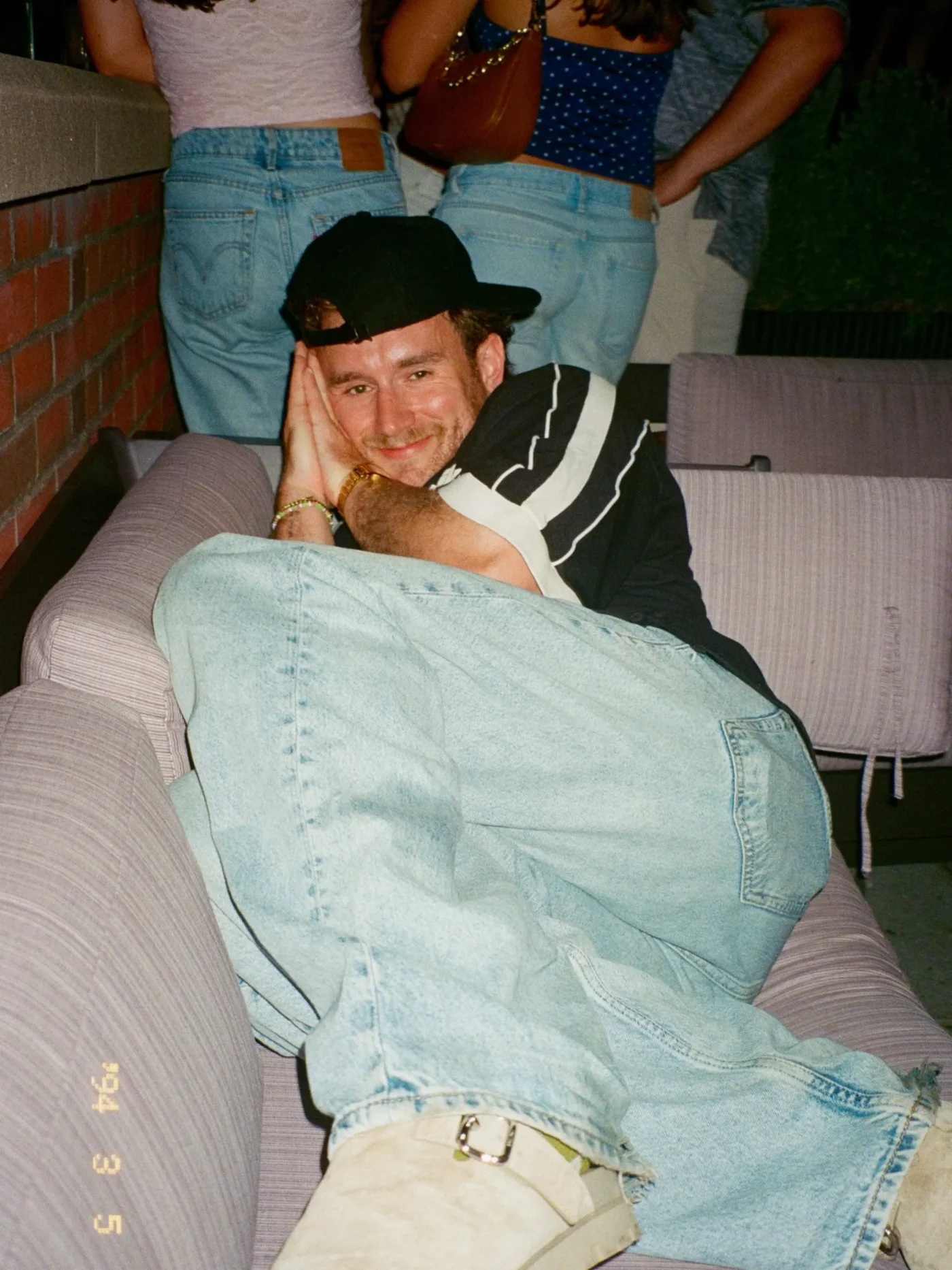 West Wilson lounging on a couch, wearing a black cap, black shirt, and light-wash jeans, with friends standing behind him.