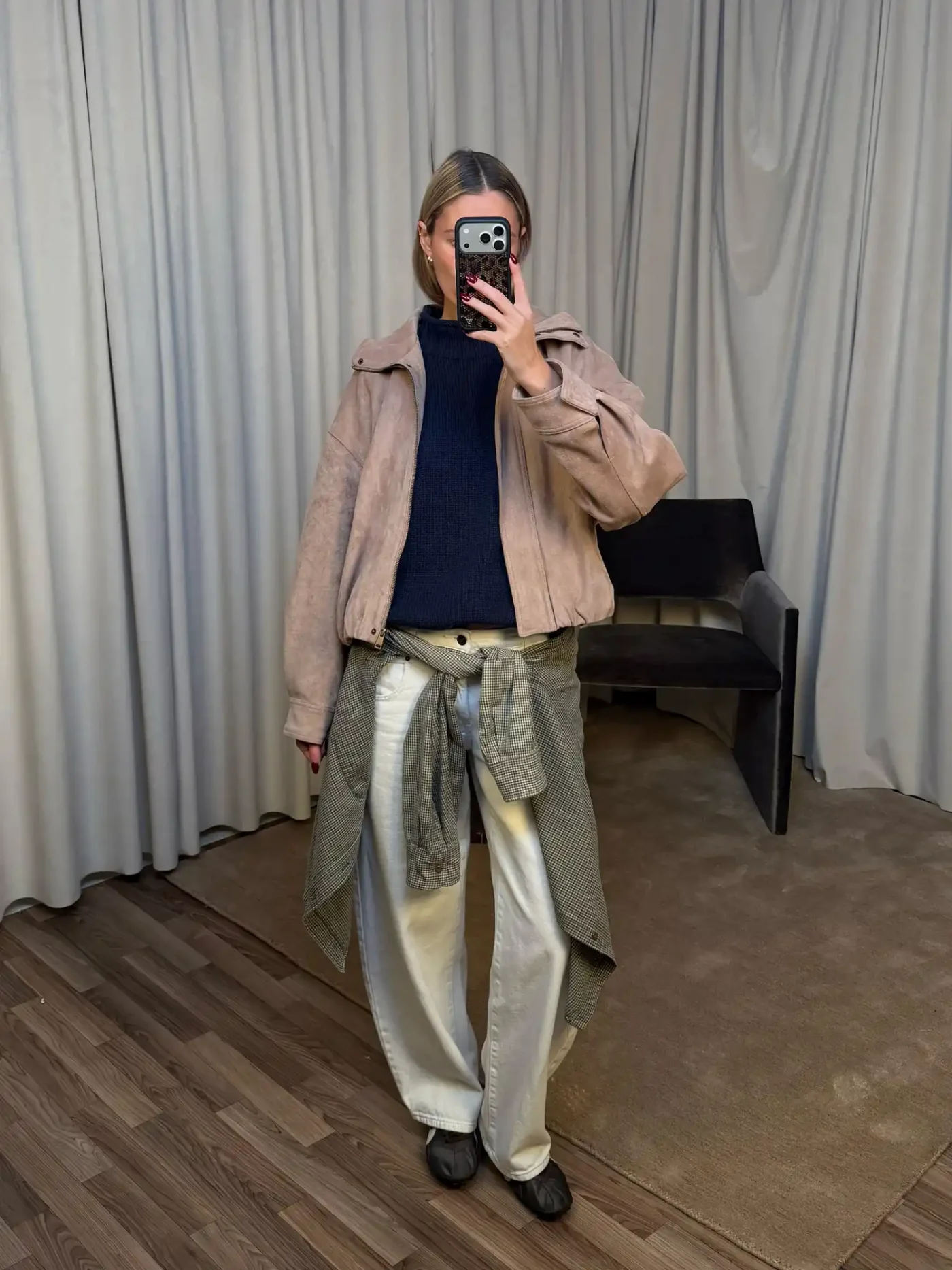 Woman taking a selfie in a mirror inside a fitting room dressed in layered casual attire with a sweater tied around her waist