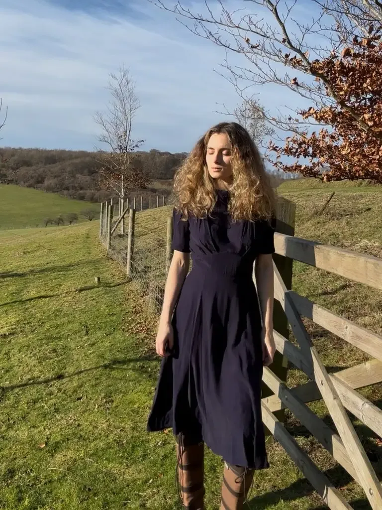 Woman with long hair in a navy dress and boots standing by a fence in a field.